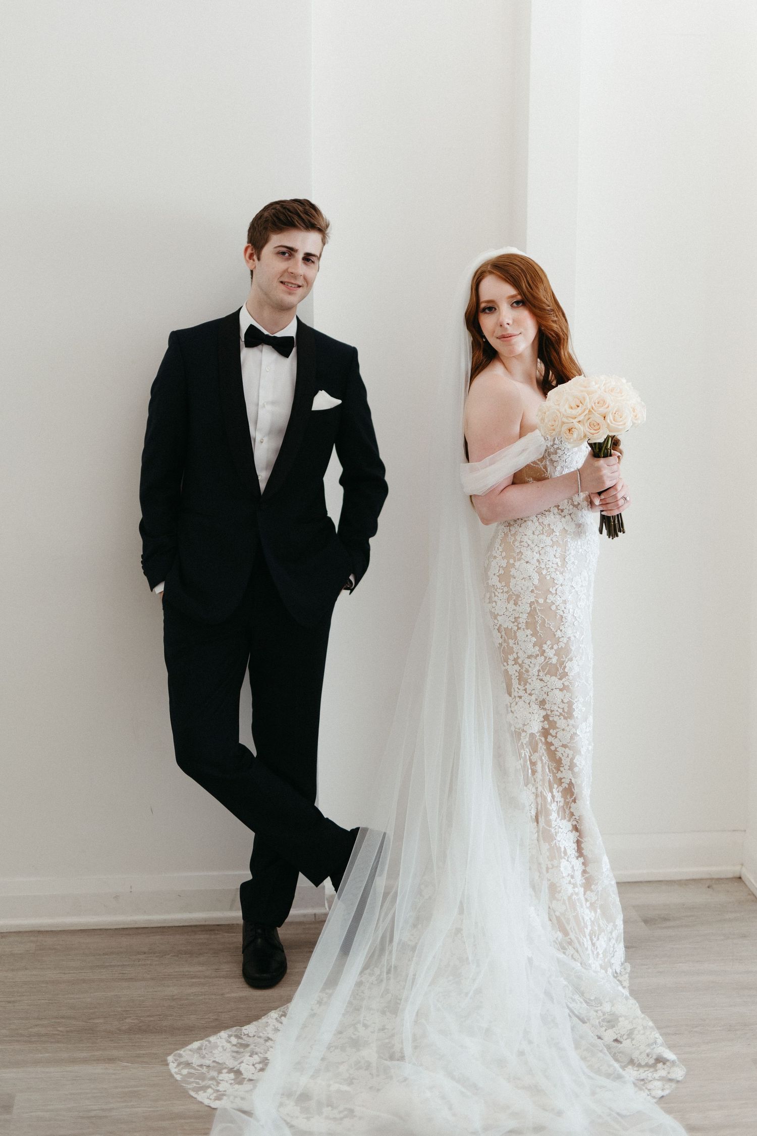 A bride in a lace gown and groom in classic tuxedo pose elegantly against a minimalist white backdrop.