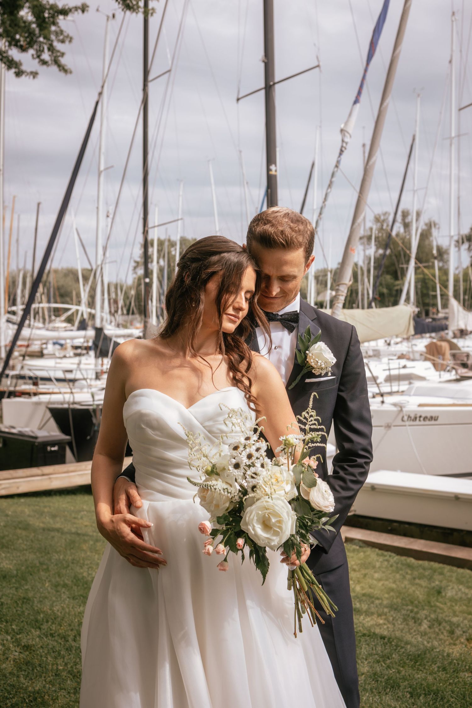 A romantic wedding photo at a marina with sailboat masts in the background and a beautiful white bouquet.
