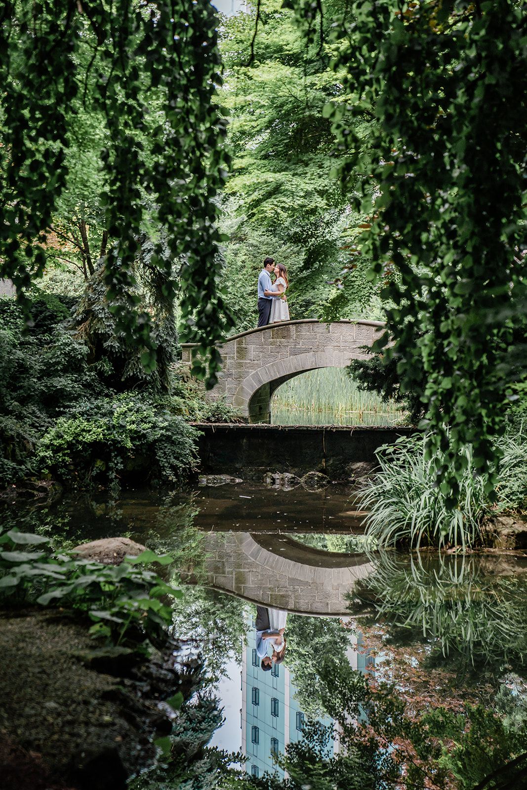 Romantic stone bridge in lush green garden with reflection in still water below.