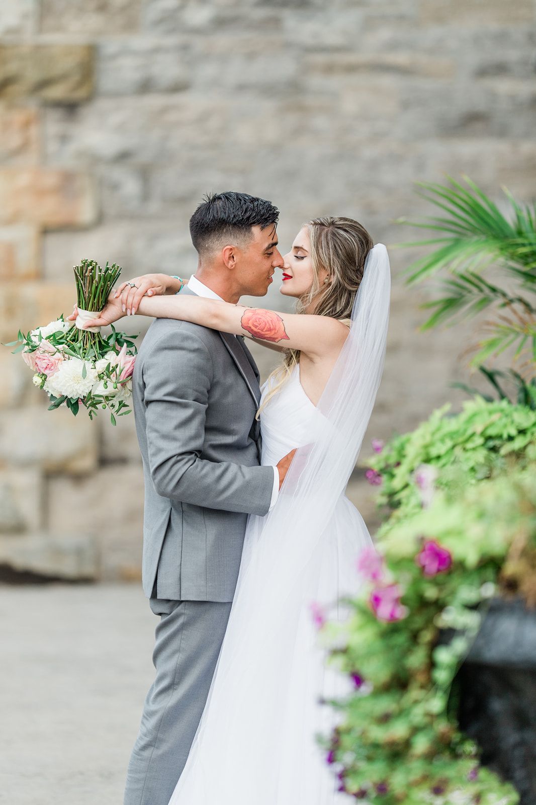 A romantic wedding photo of a couple embracing against a stone wall with flowers and greenery in the background.