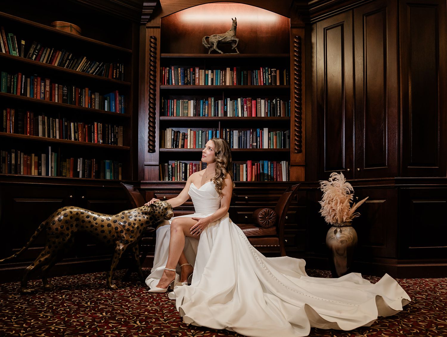 Bride in flowing white gown poses in elegant library setting with dark wood bookshelves.