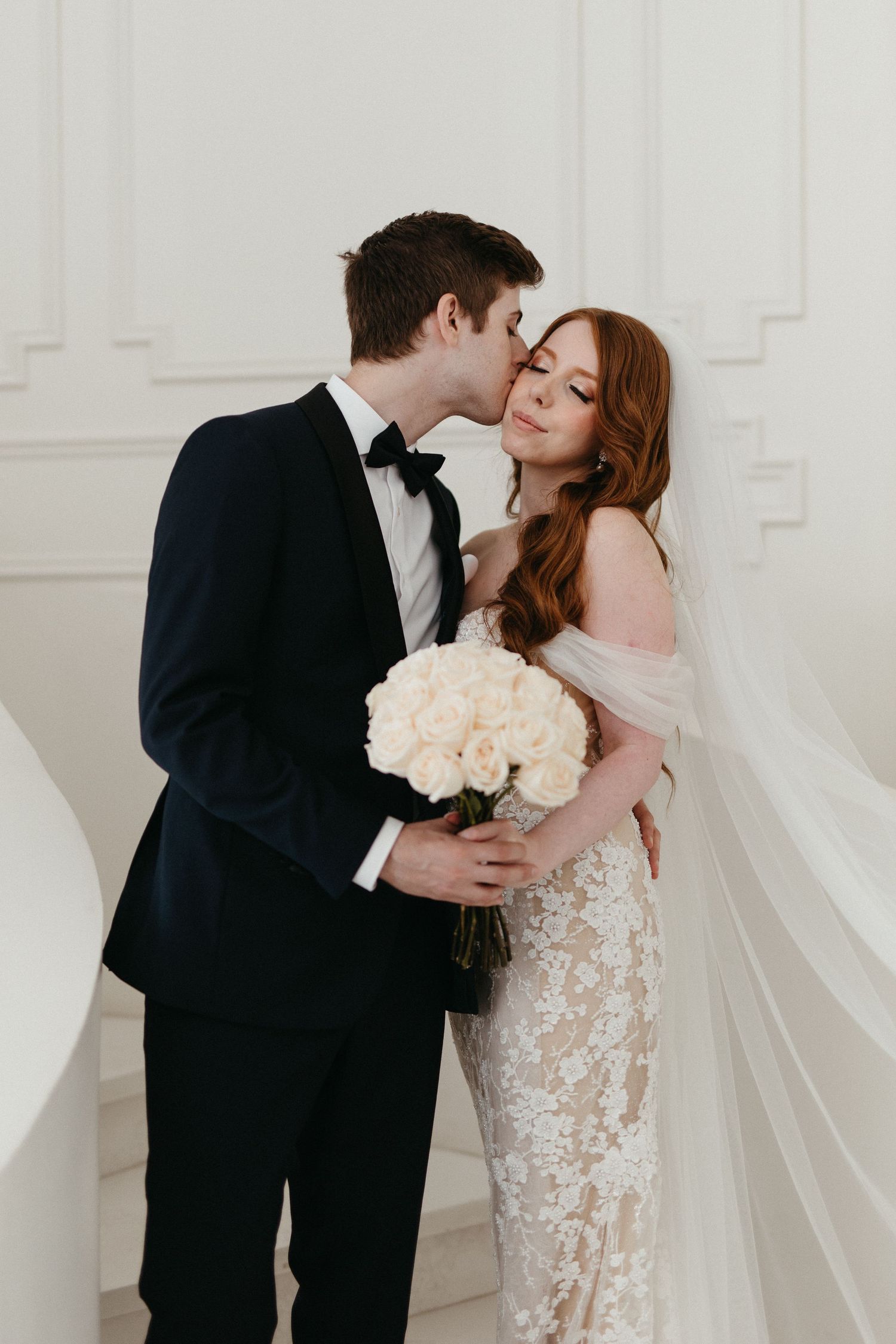 Romantic wedding moment captured in white room with couple holding white rose bouquet and sharing tender kiss.