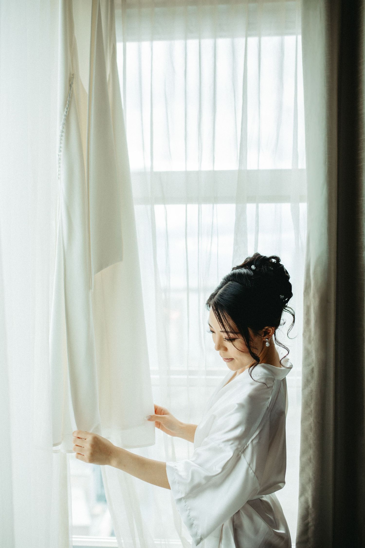 A figure in a white silk robe stands by a window with sheer curtains in soft morning light.