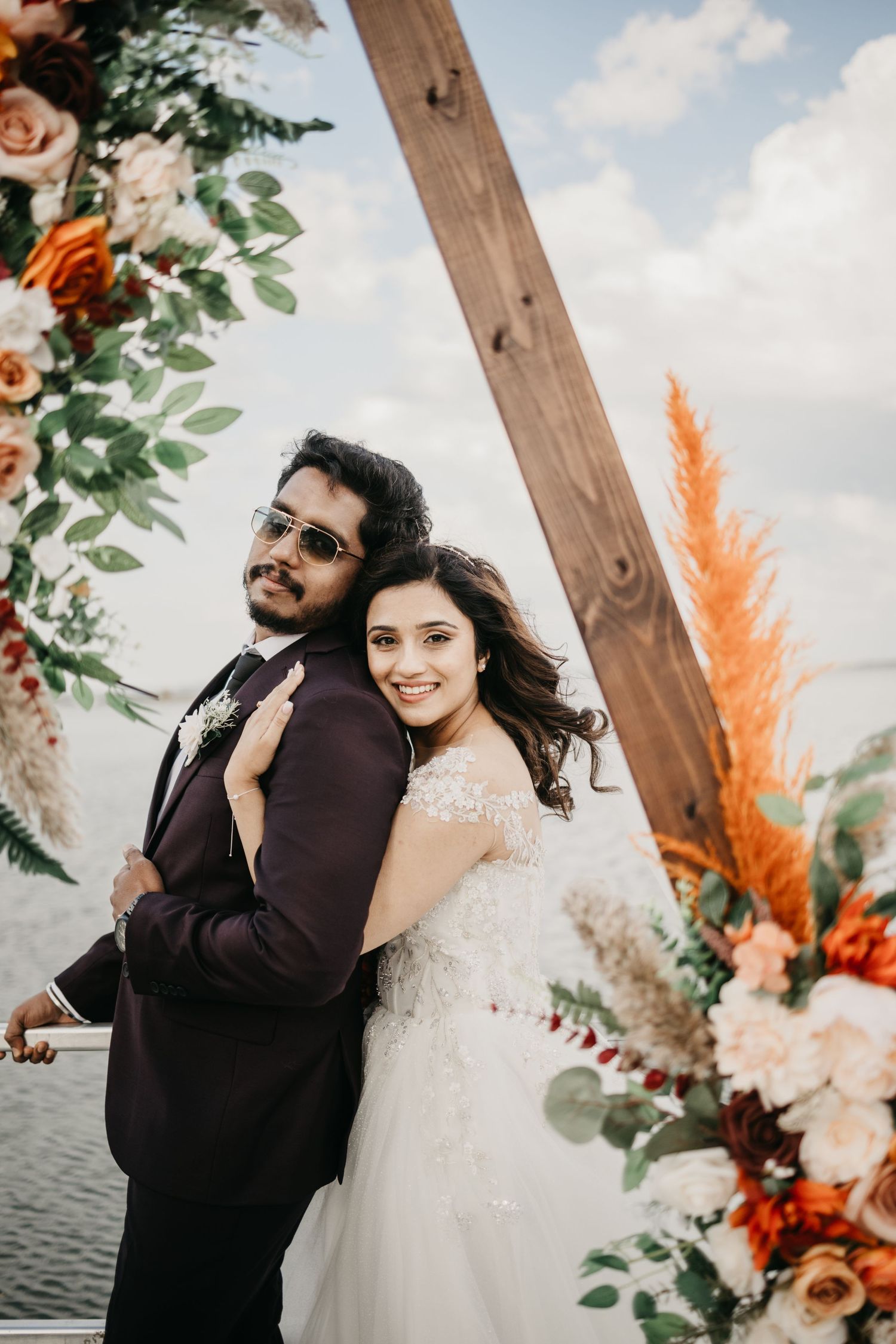 A couple poses beneath a wooden arch decorated with roses, eucalyptus, and pampas grass against a cloudy sky.