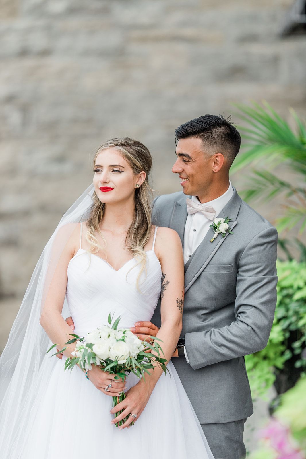Elegant newlyweds pose together outdoors with white bouquet next to a stone wall and tropical greenery.
