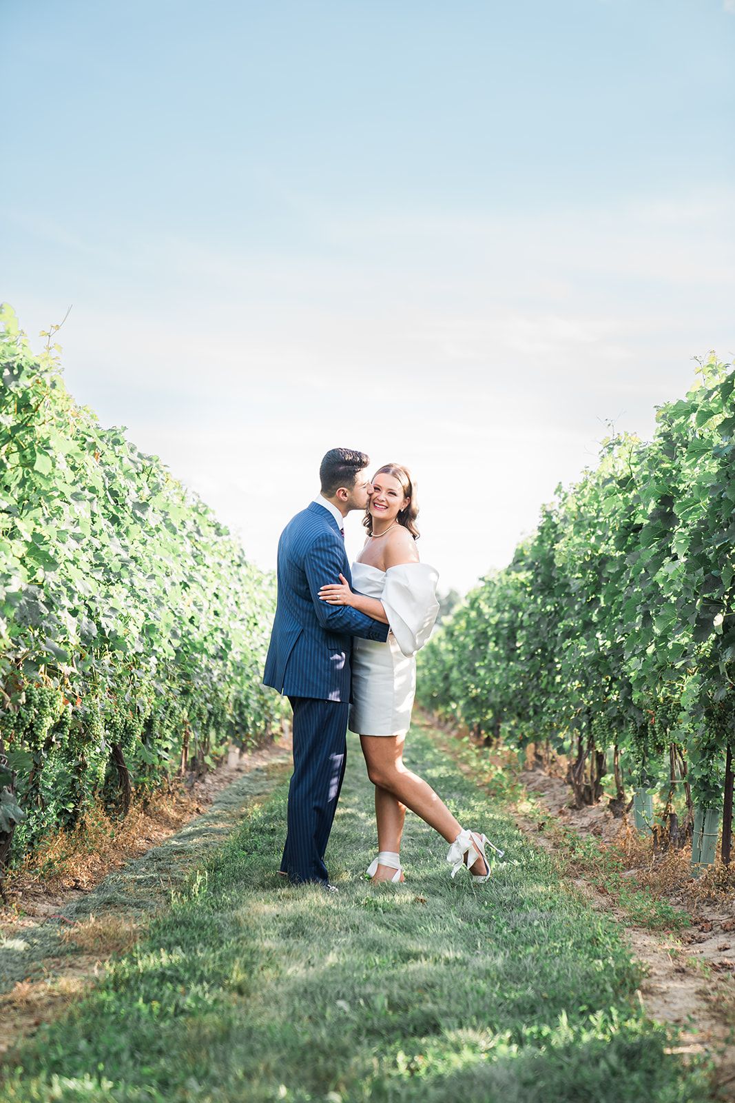 Couple embracing in a lush vineyard with bright blue sky overhead.