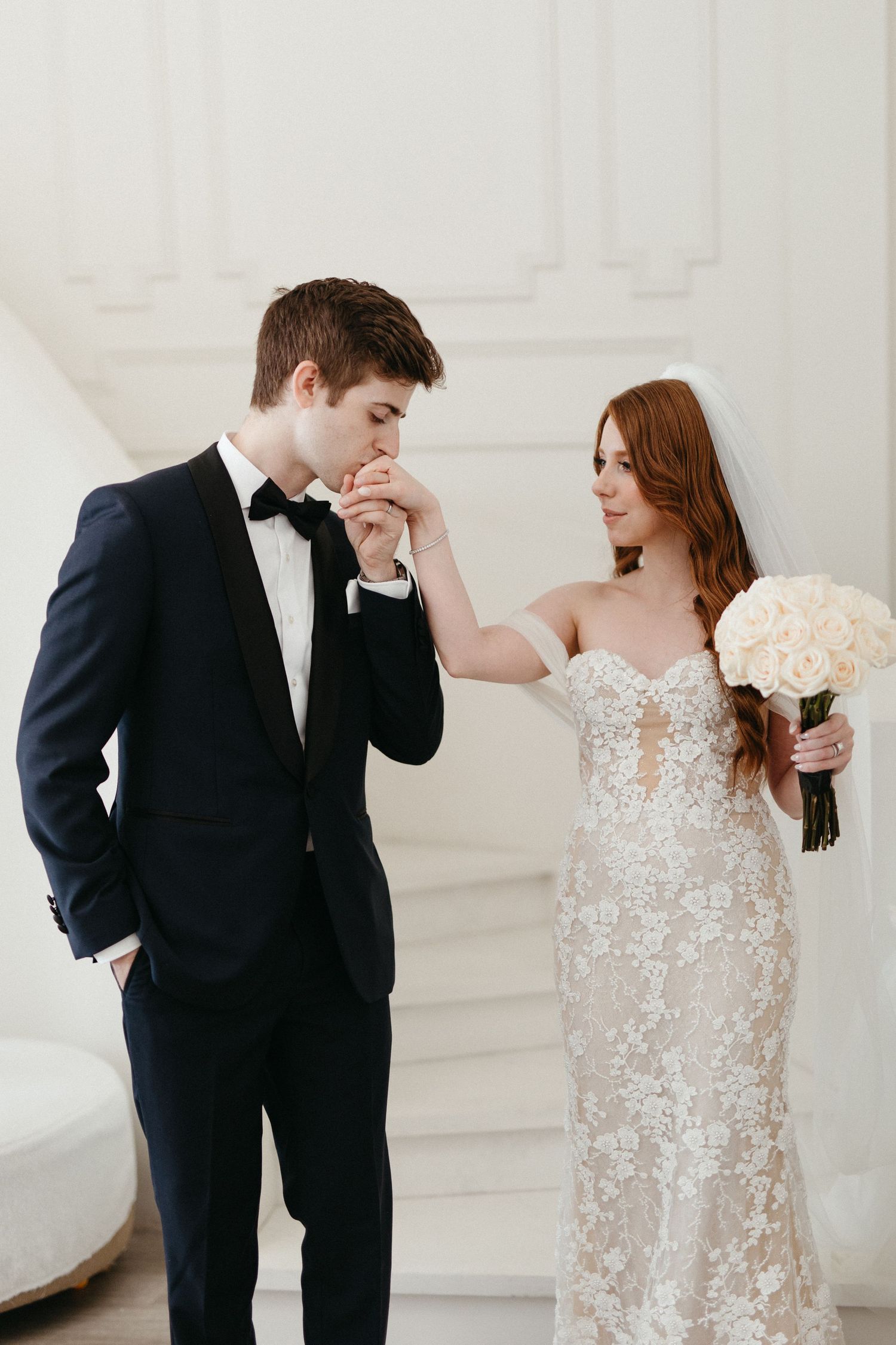 A couple in wedding attire share an intimate moment with the bride holding a white bouquet against a white wall.