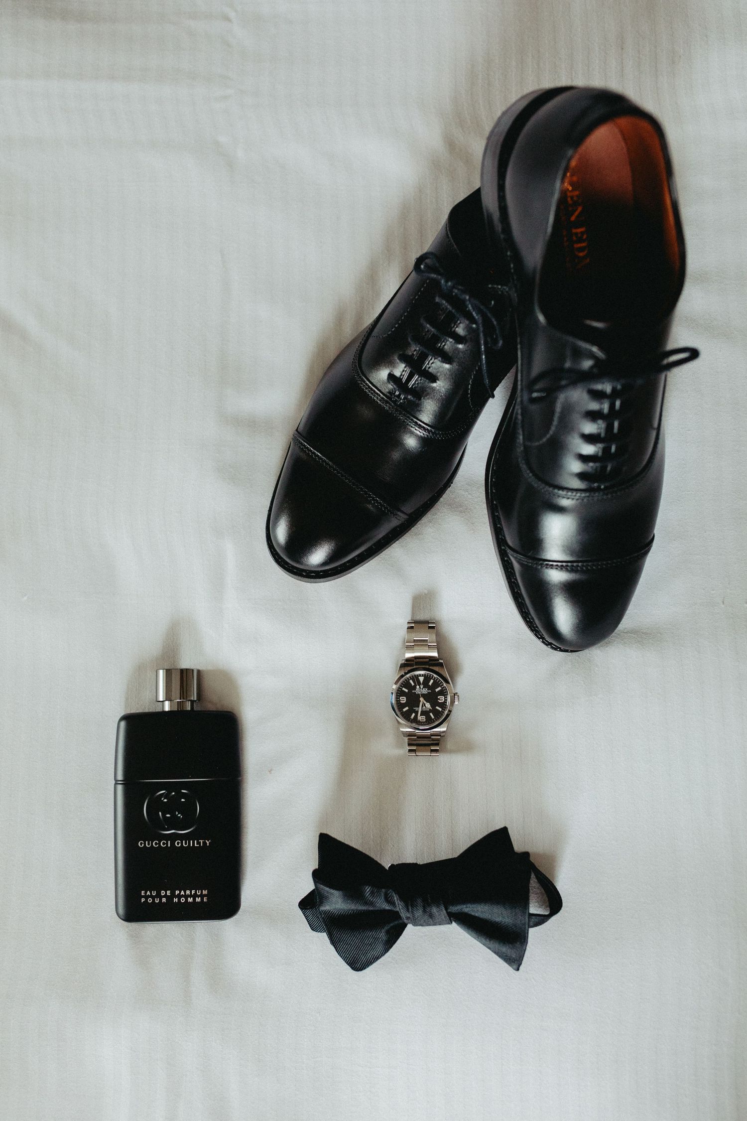 A flat lay of men's formal accessories including black oxford shoes, a wristwatch, black bow tie, and cologne on white fabric.