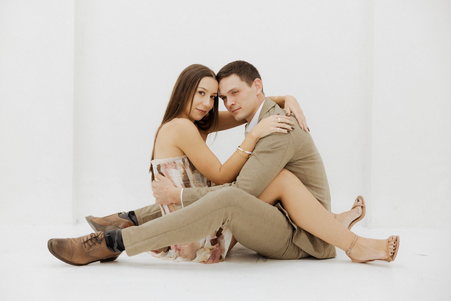 Two people in neutral colors embrace while sitting together on a white backdrop.