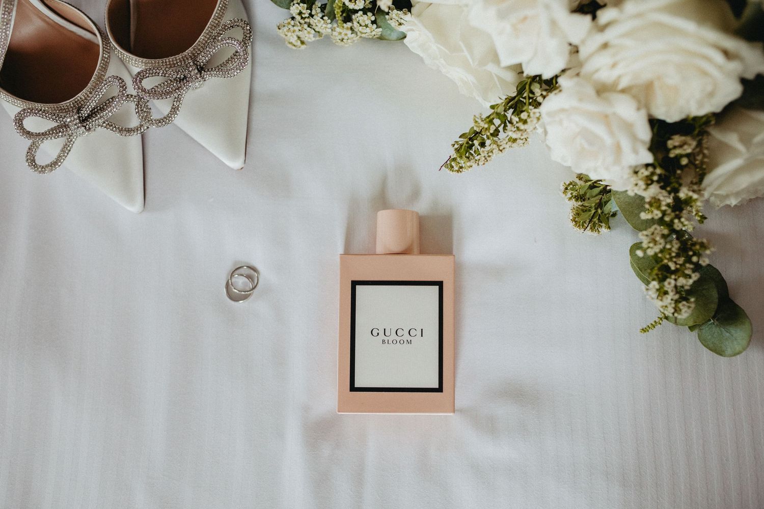 Wedding details with a Gucci perfume bottle, silver shoes, and delicate white floral arrangements on a white surface.