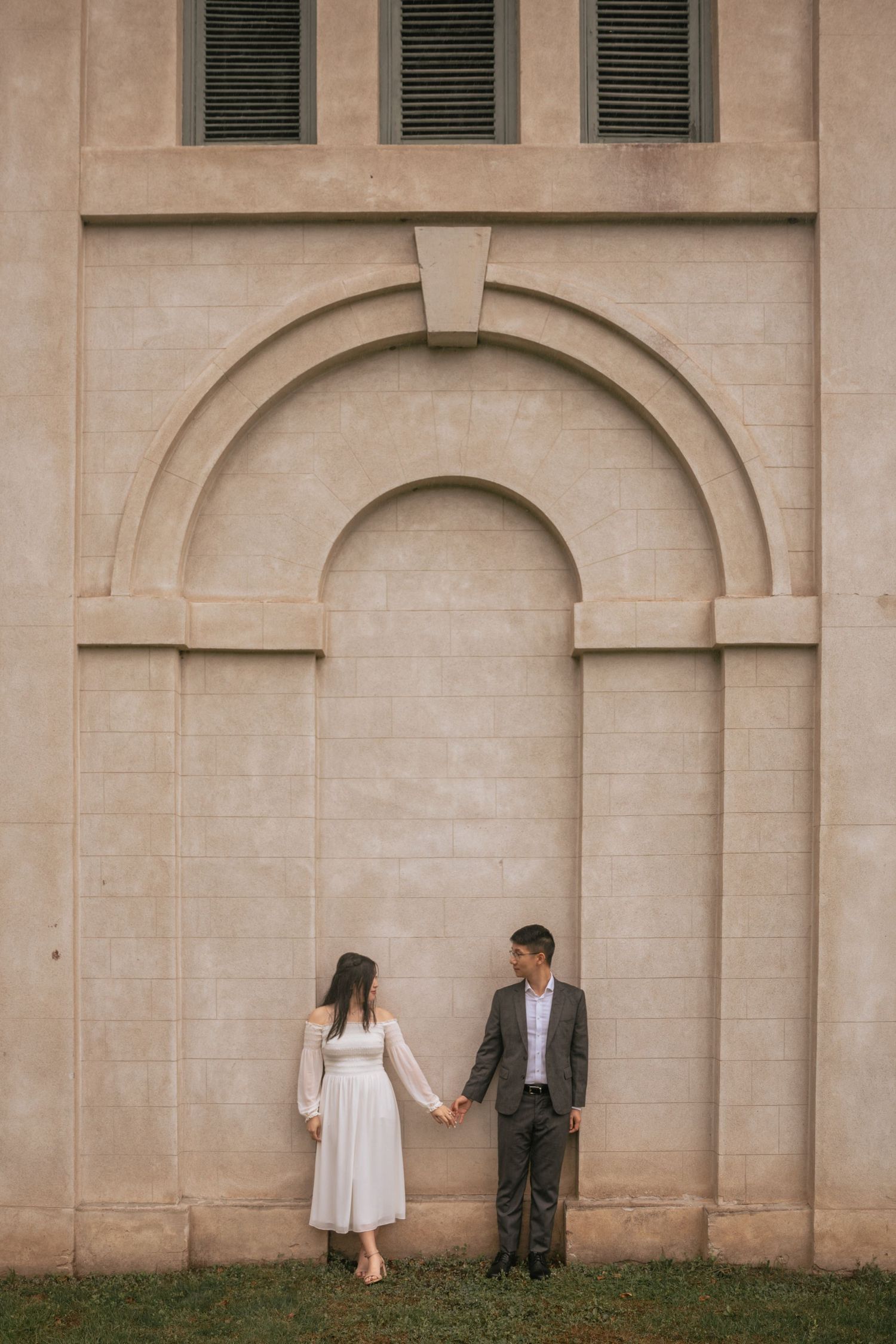 A couple stands holding hands against a beige stone wall with arched architectural details and green shutters.