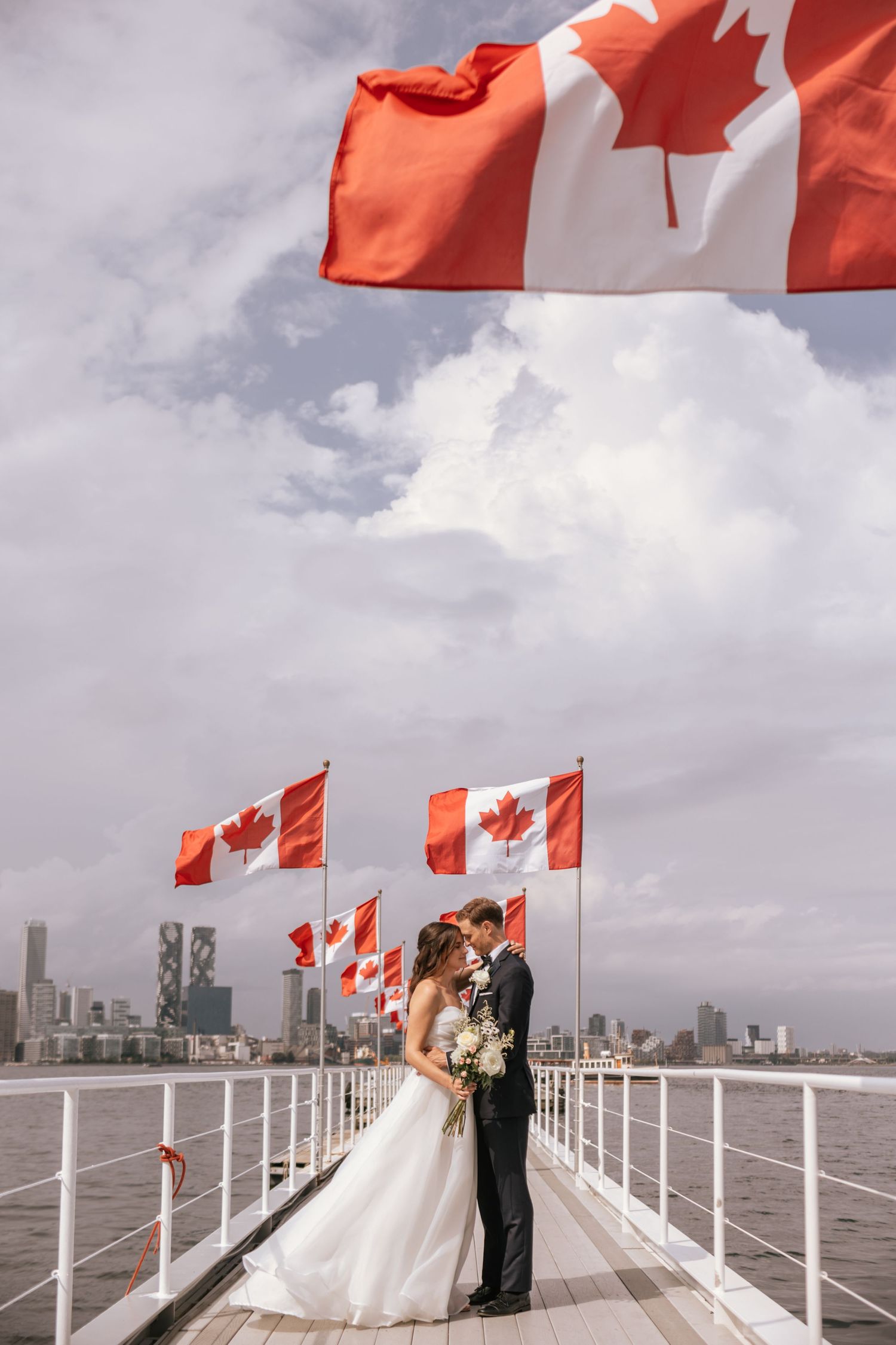 Canadian flag waves over couple on pier with Toronto skyline in background under cloudy skies.