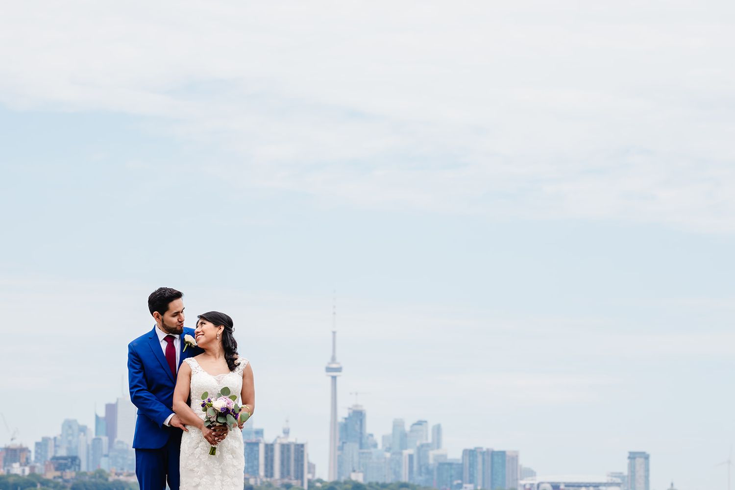 Wedding couple stands against Toronto skyline with CN Tower visible in background on sunny day.