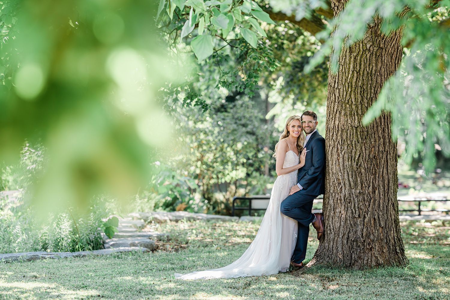 A couple shares a romantic moment under a large tree in a dreamy outdoor garden setting with soft bokeh lighting effects.