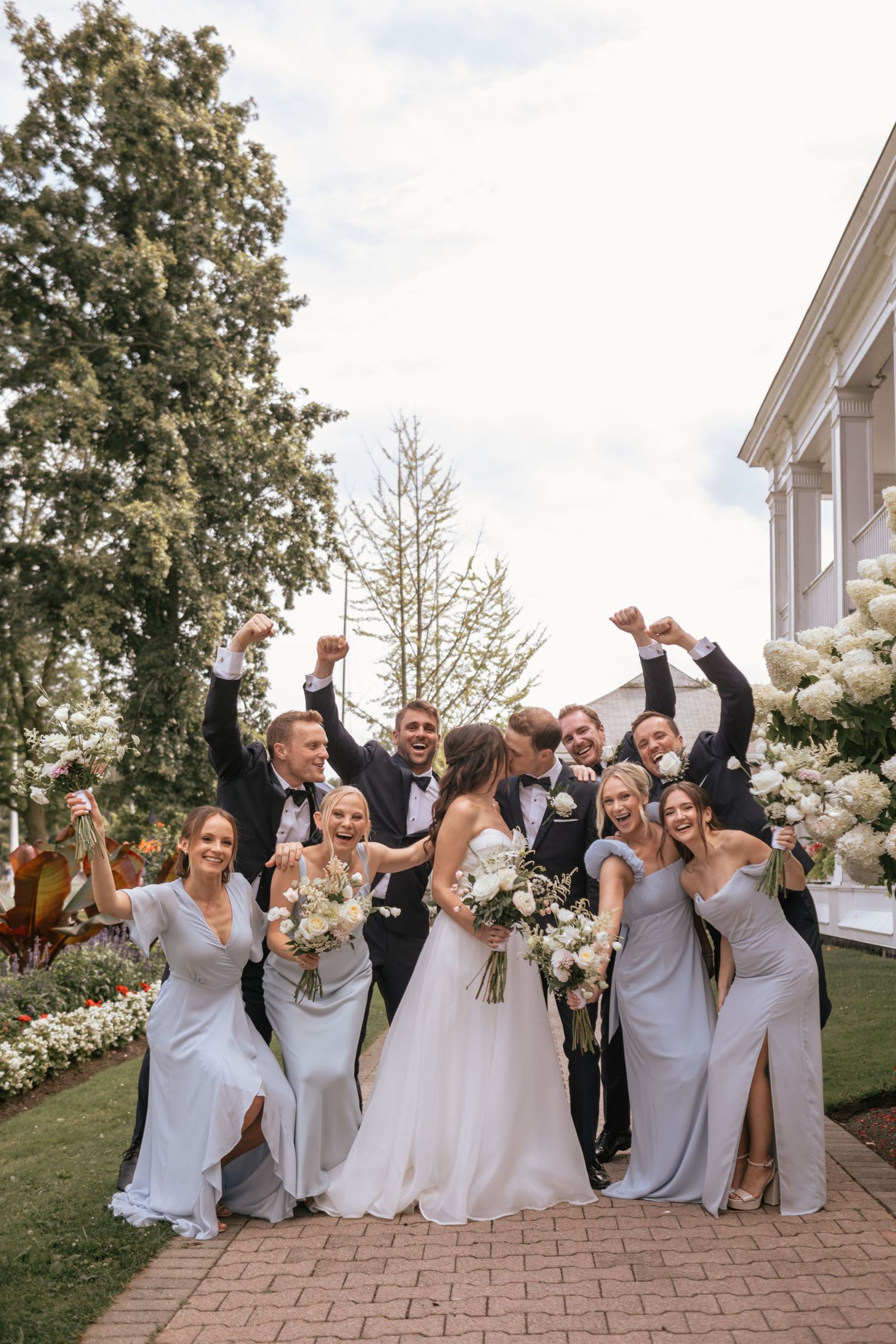 Wedding party celebrating with raised arms on a landscaped lawn with white floral decorations.