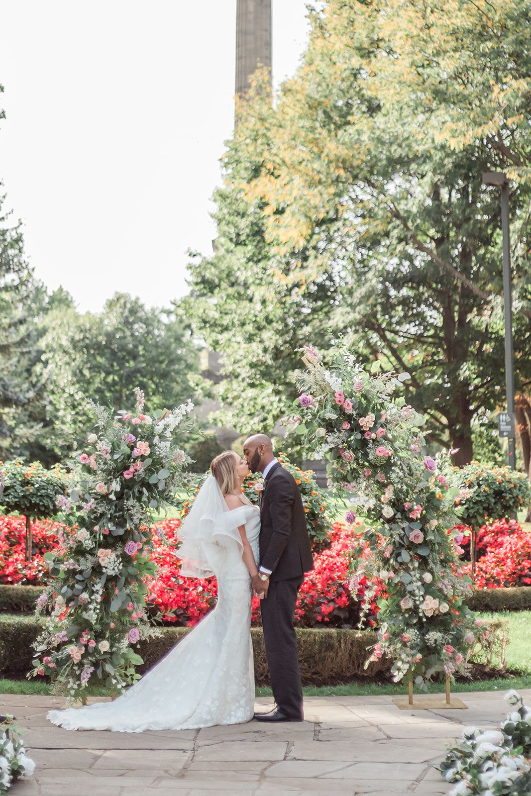 Wedding couple embraces under a floral arch with pink roses and red azaleas in a lush garden setting.