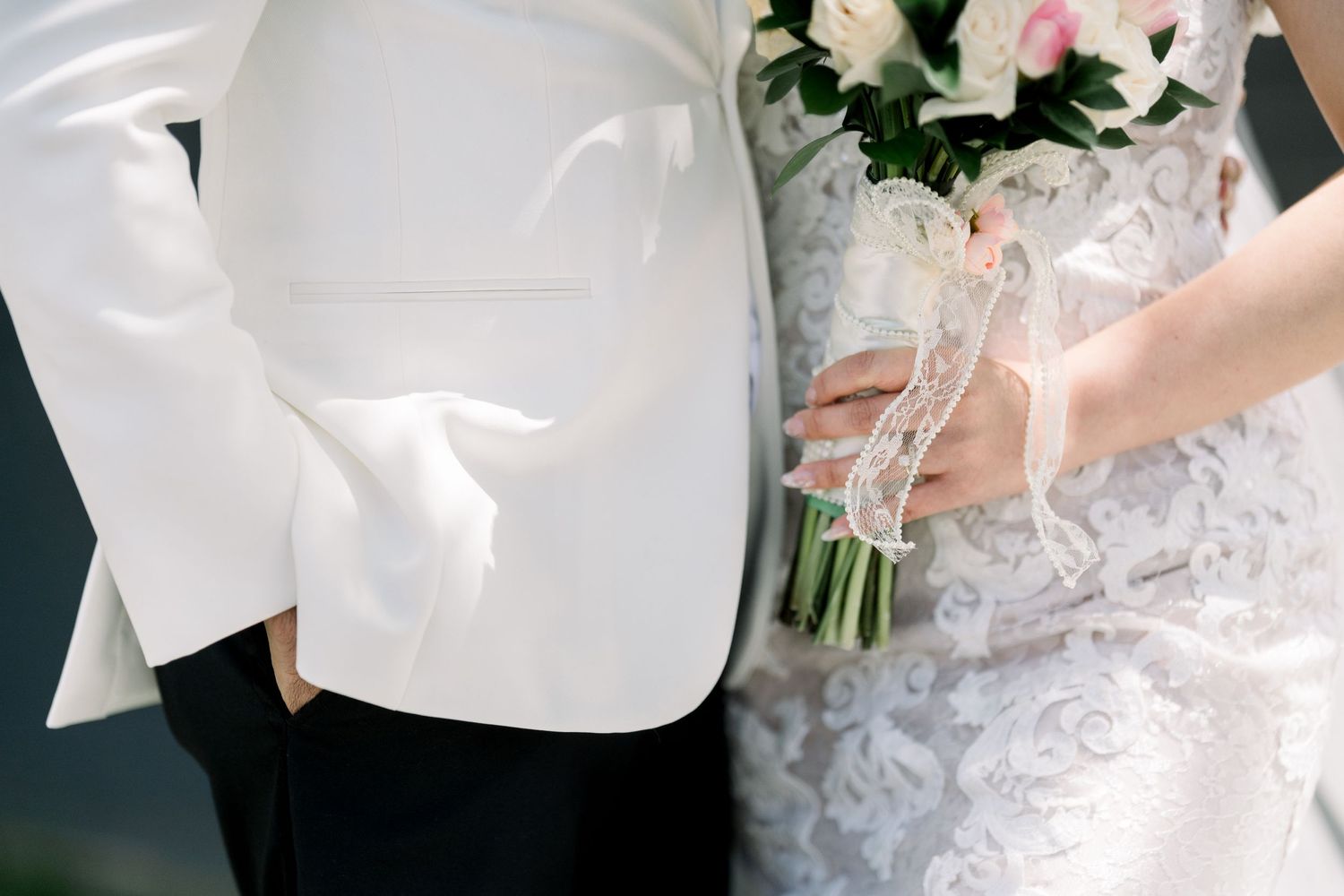 Detail shot of white tuxedo jacket and lace wedding dress with pink and white bouquet.