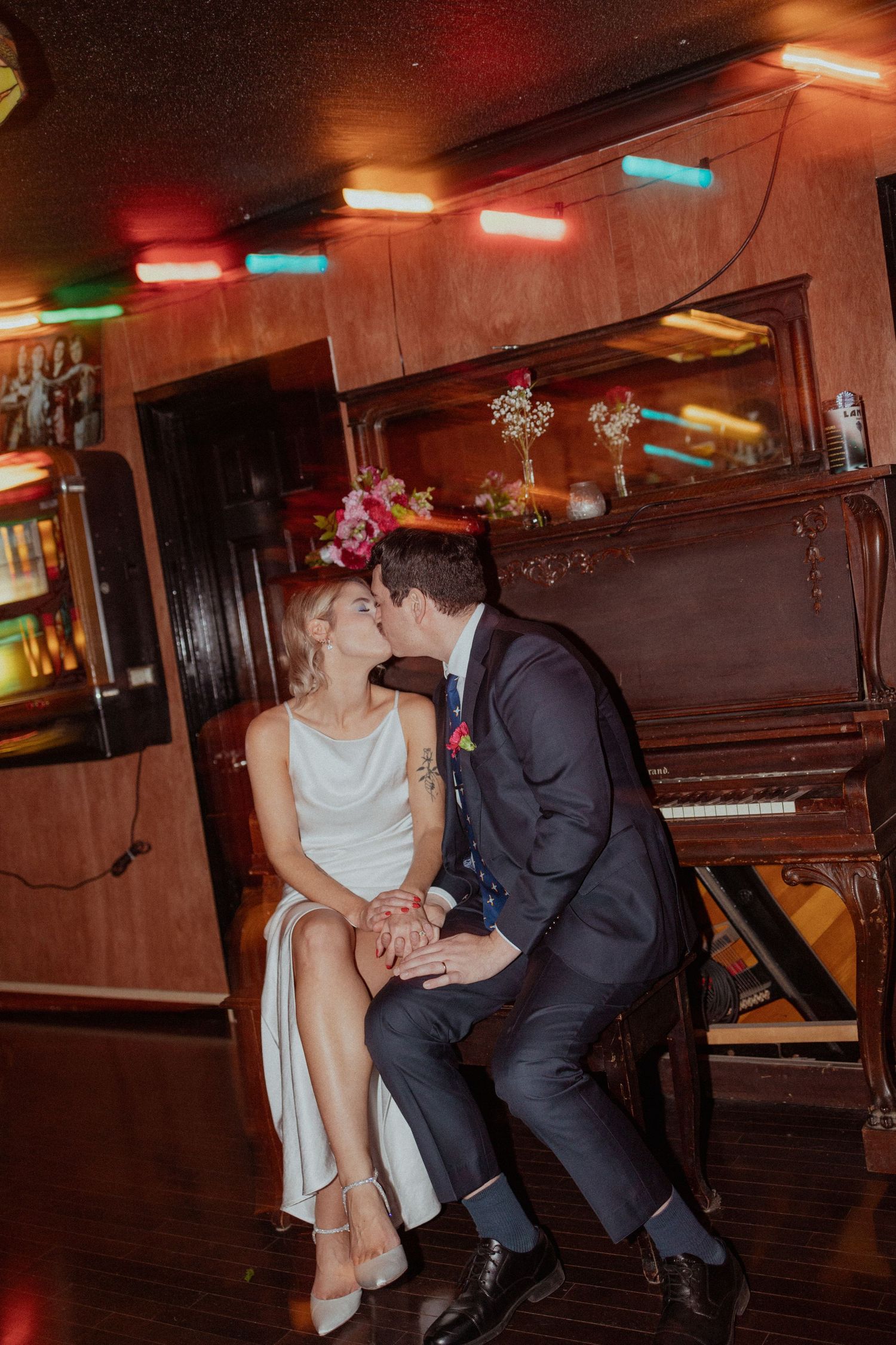Romantic couple in evening attire share an intimate moment by an upright piano under colorful bar lighting.