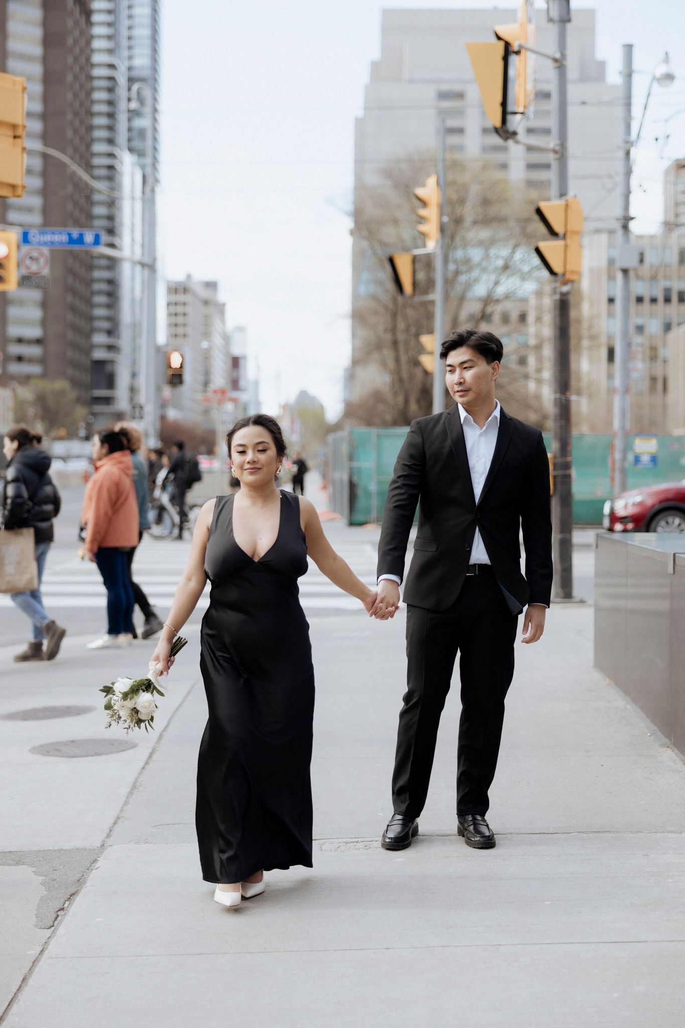 A couple in elegant black attire walks hand in hand on a city sidewalk.