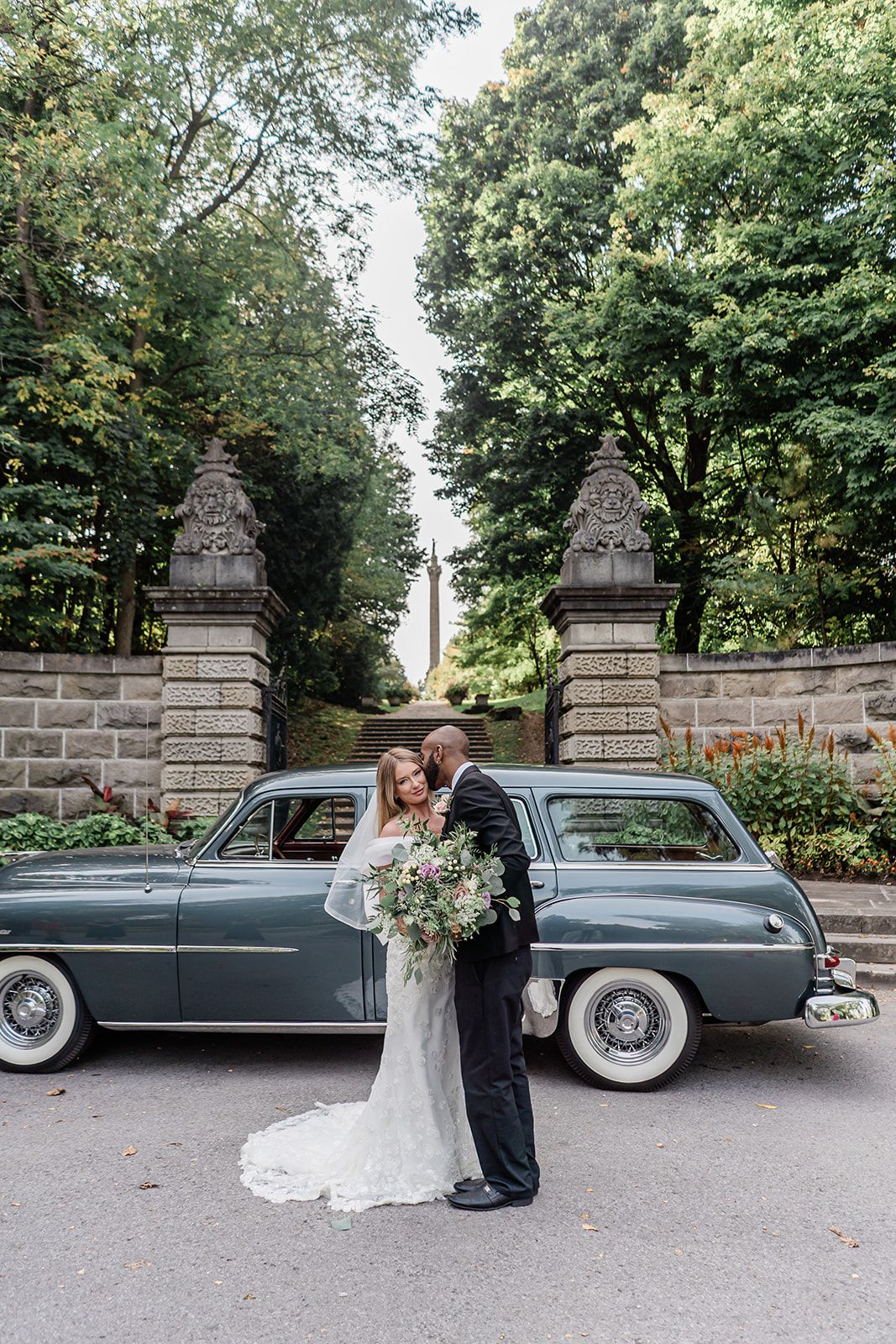 Classic vintage car parked between stone pillars at elegant garden entrance with couple in wedding attire.