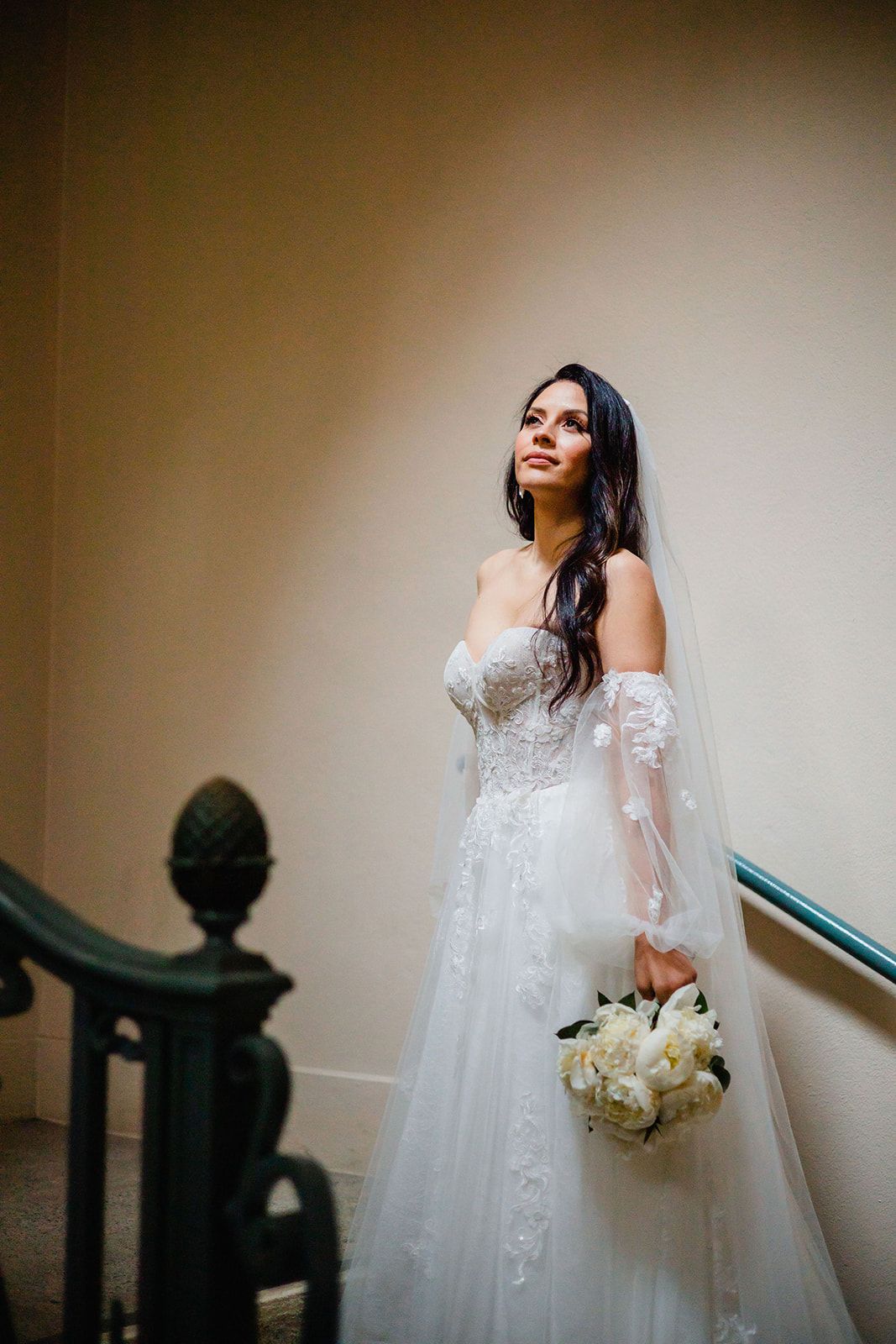 Bride in white lace wedding gown holds bouquet while standing on elegant staircase.