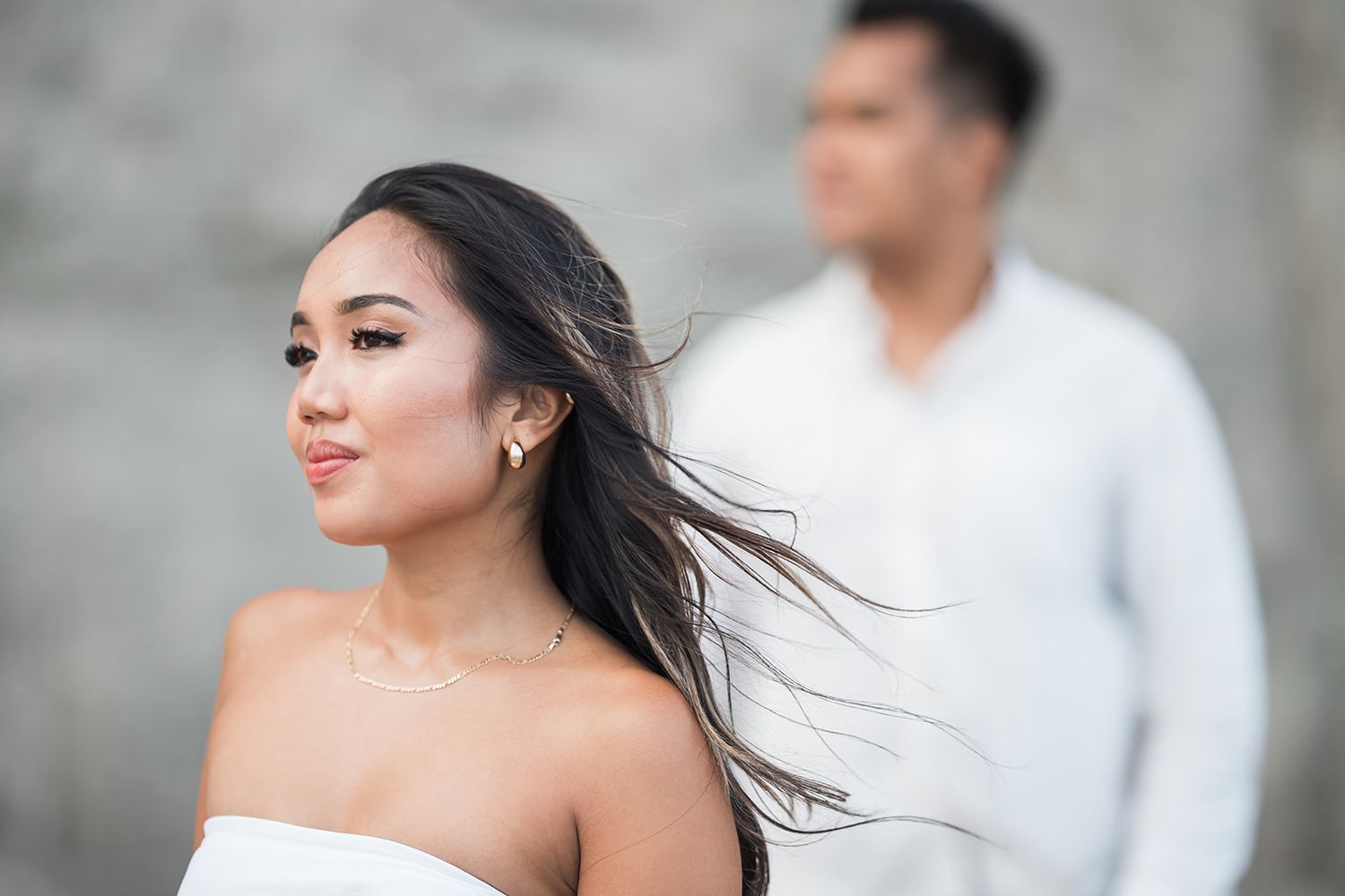 A person in a white off-shoulder top with long dark hair flowing in the breeze, with a blurred figure in white behind.