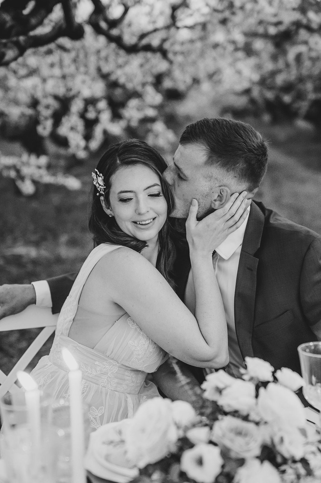 A romantic black and white wedding photo showing a tender moment between newlyweds at an outdoor reception with spring blossoms.