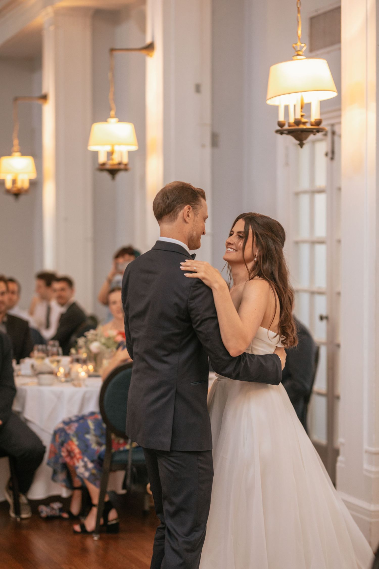 A couple shares their first dance at an elegant wedding reception with warm lighting and ornate wall sconces.