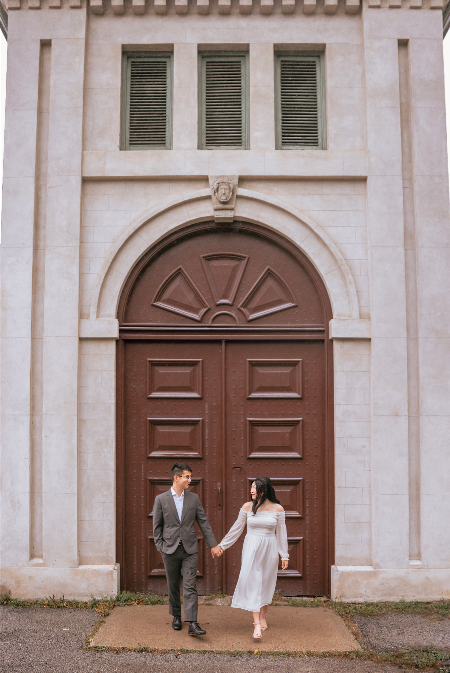 A couple stands in front of a large arched brown door with green shutters on a white building exterior.