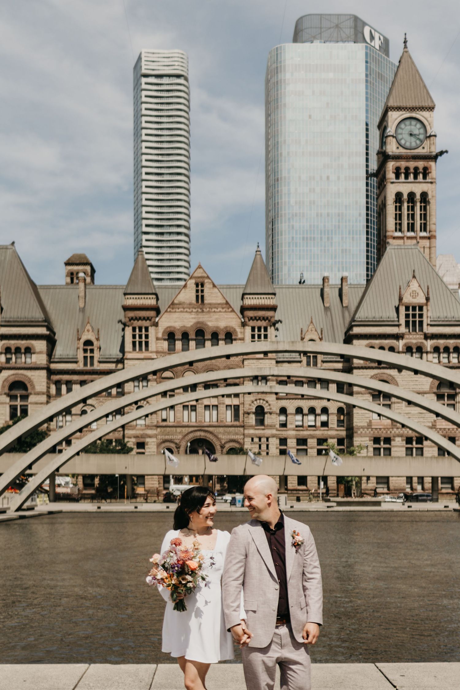 A couple stands in front of Toronto's Old City Hall with modern skyscrapers rising behind the historic architecture.