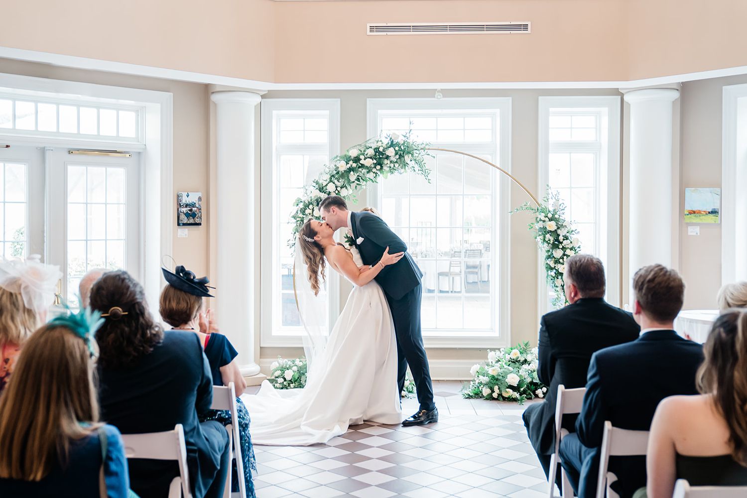 Wedding ceremony in a bright sunlit room with a floral arch and seated guests watching the celebration.