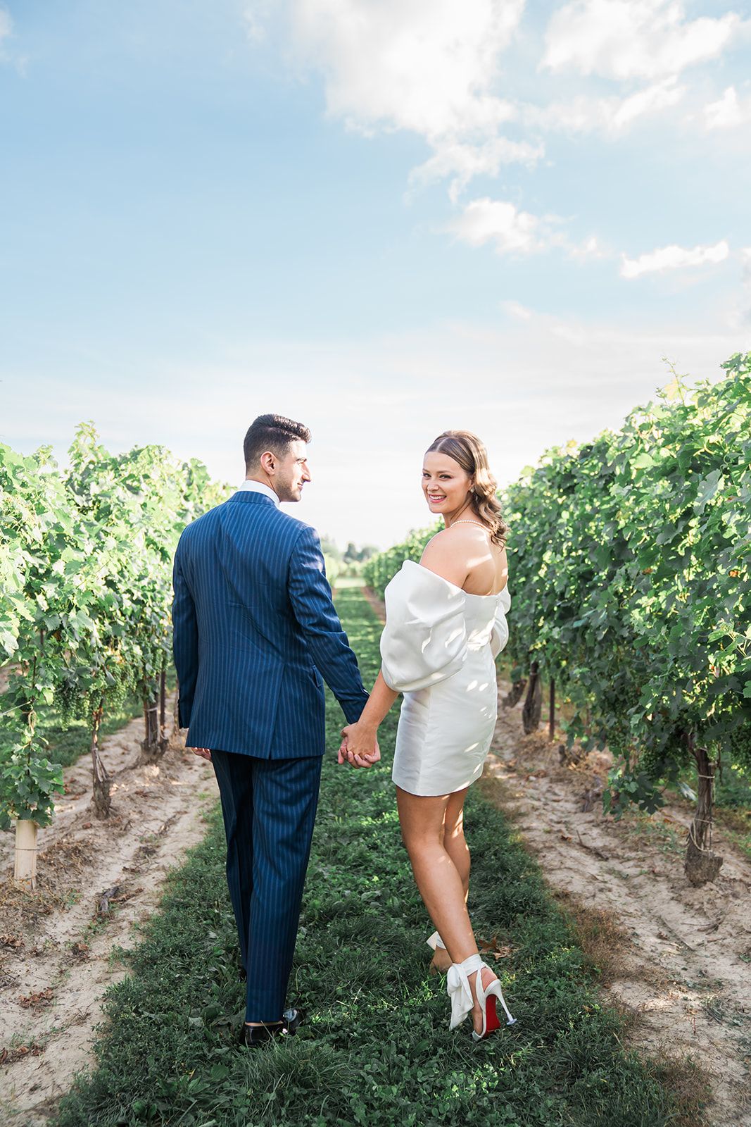 Couple holding hands while walking between vineyard rows on a sunny day with blue skies and green vines.