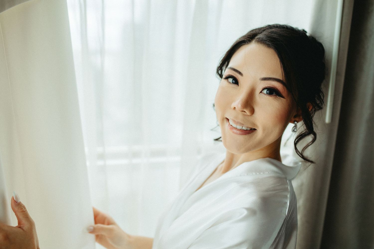 Smiling woman in white robe stands by sheer curtain window during morning preparations.