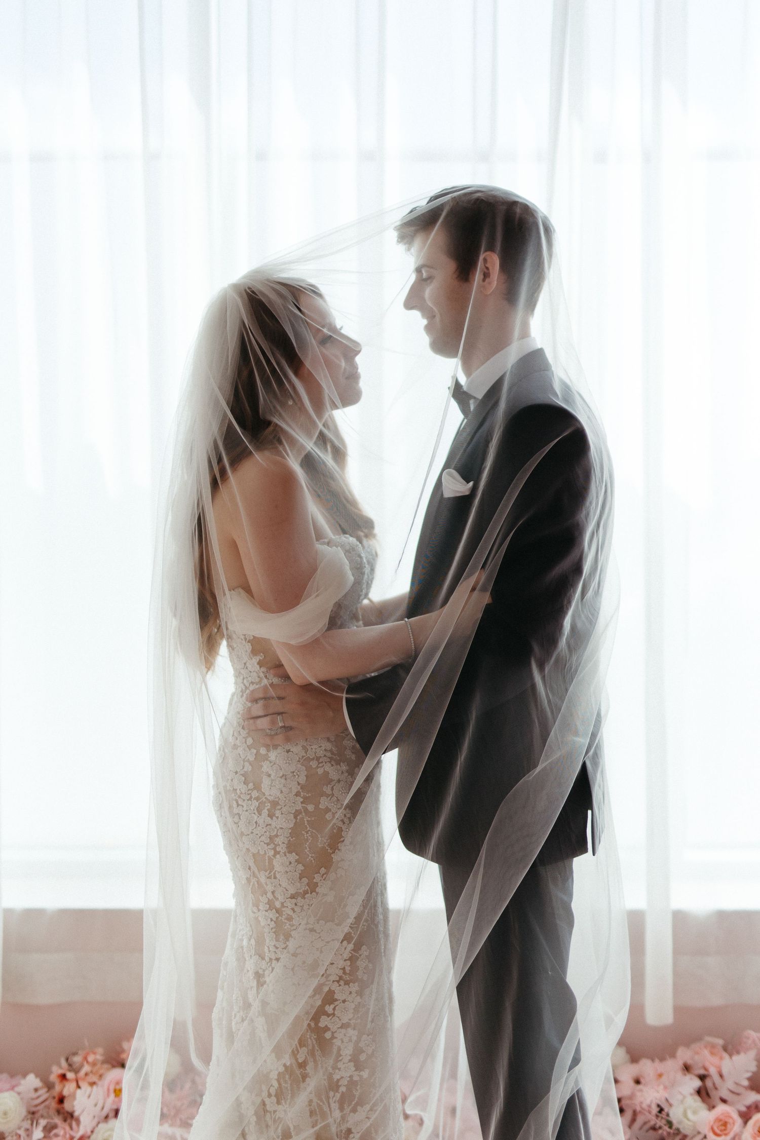 A romantic wedding portrait silhouetted against sheer white curtains with pink floral decor at their feet.