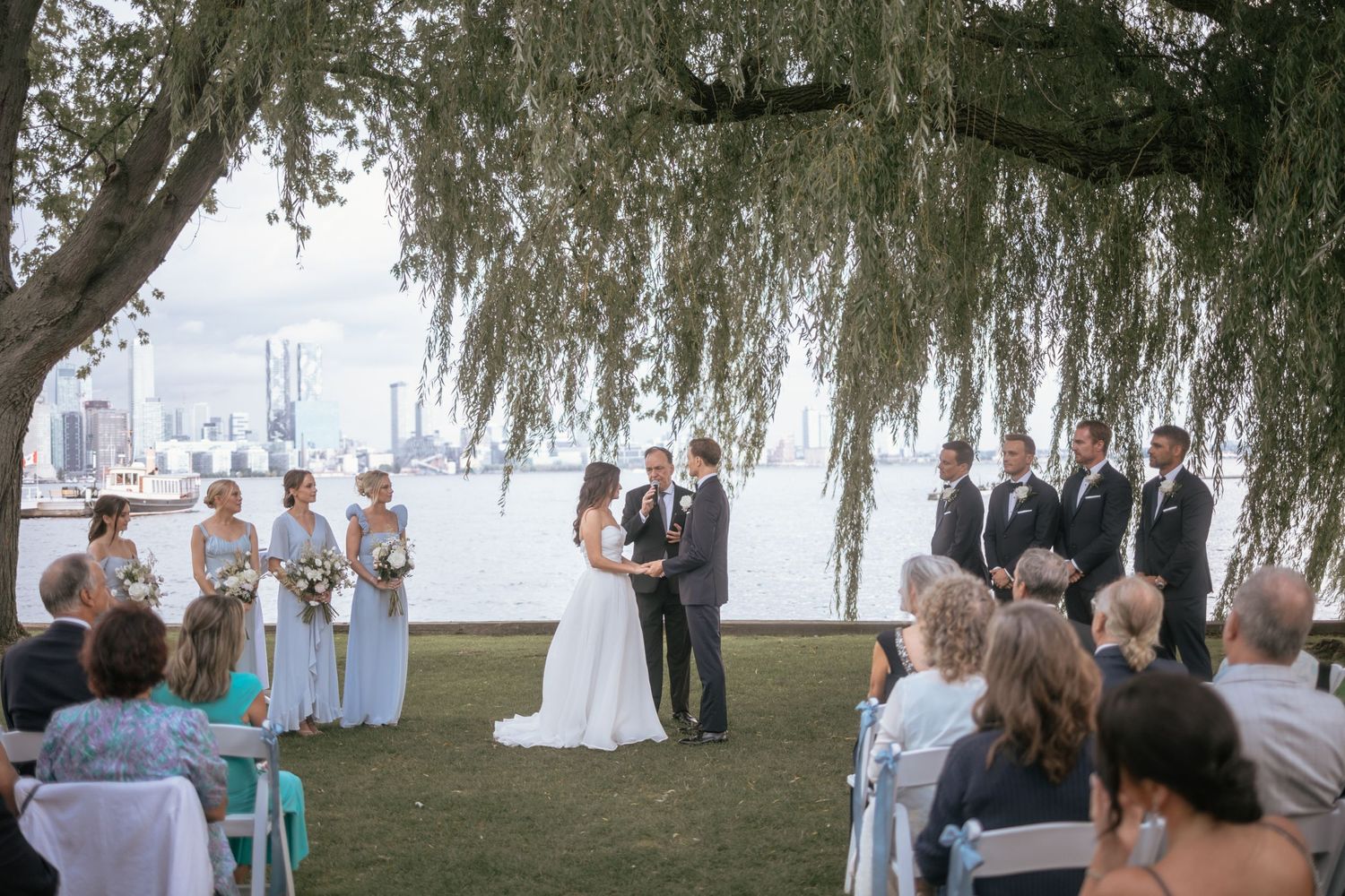 A wedding ceremony takes place under a sweeping willow tree by the water with guests seated on white chairs.