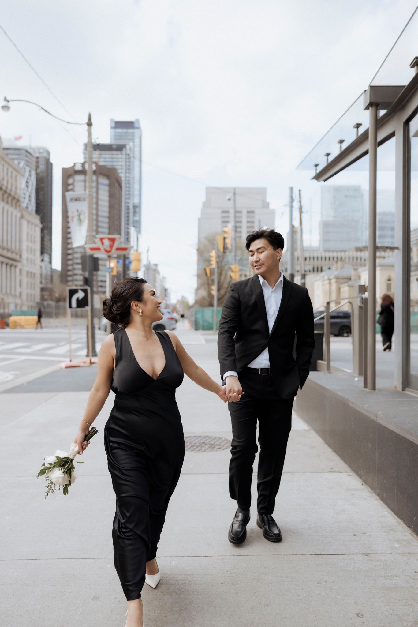 A couple walks hand in hand on an urban sidewalk with city buildings and a small bouquet.