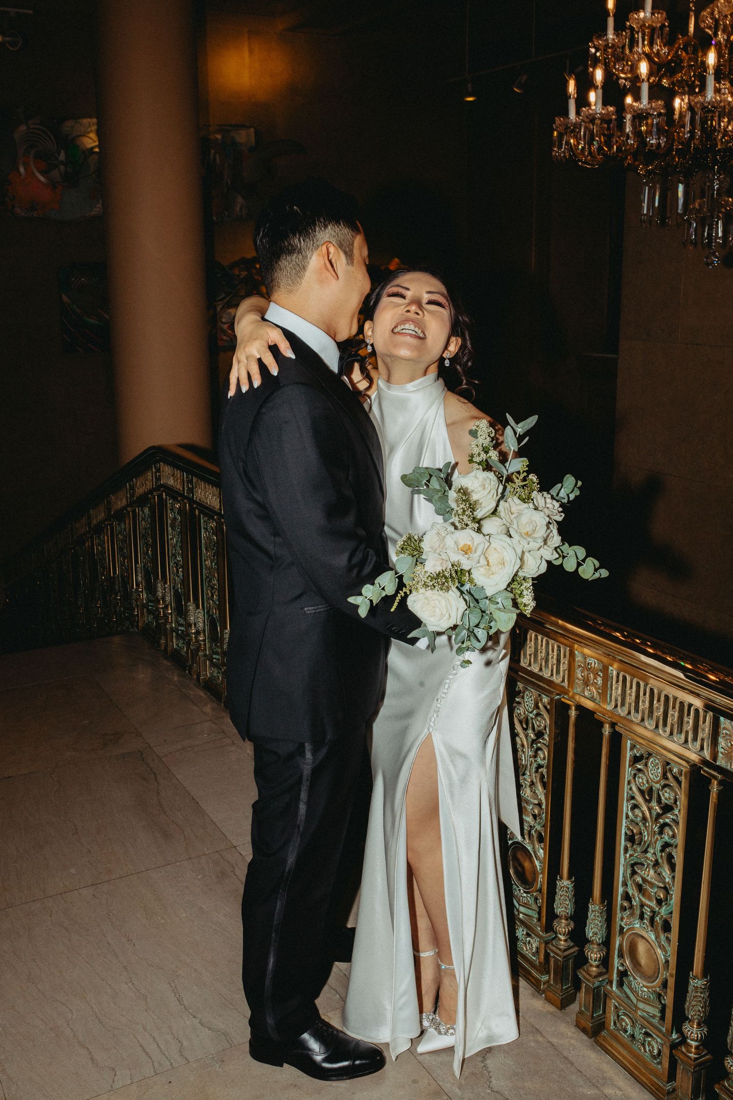 A romantic wedding moment captured in an elegant venue with a crystal chandelier and ornate railing.