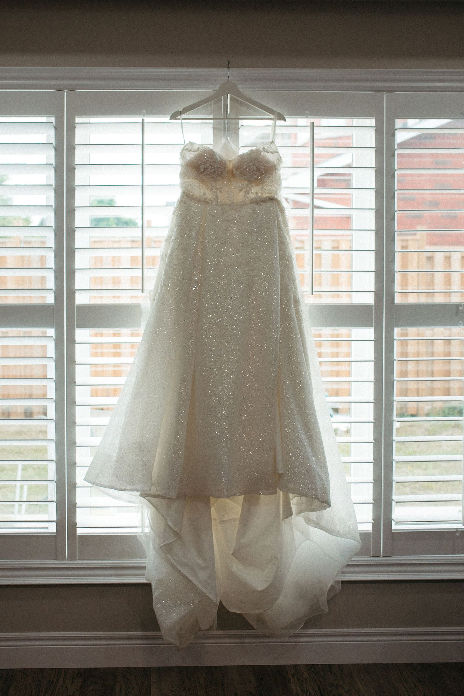 A white lace wedding dress hangs elegantly in front of a window with white shutters.