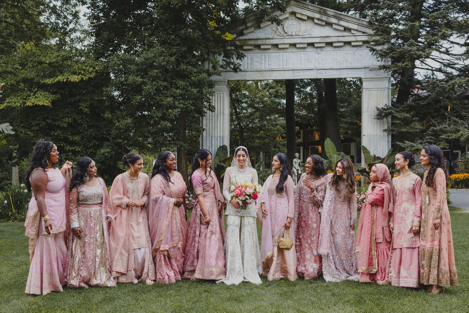 Bridal party wearing pink and blush-colored dresses pose together under a white garden arch surrounded by greenery.