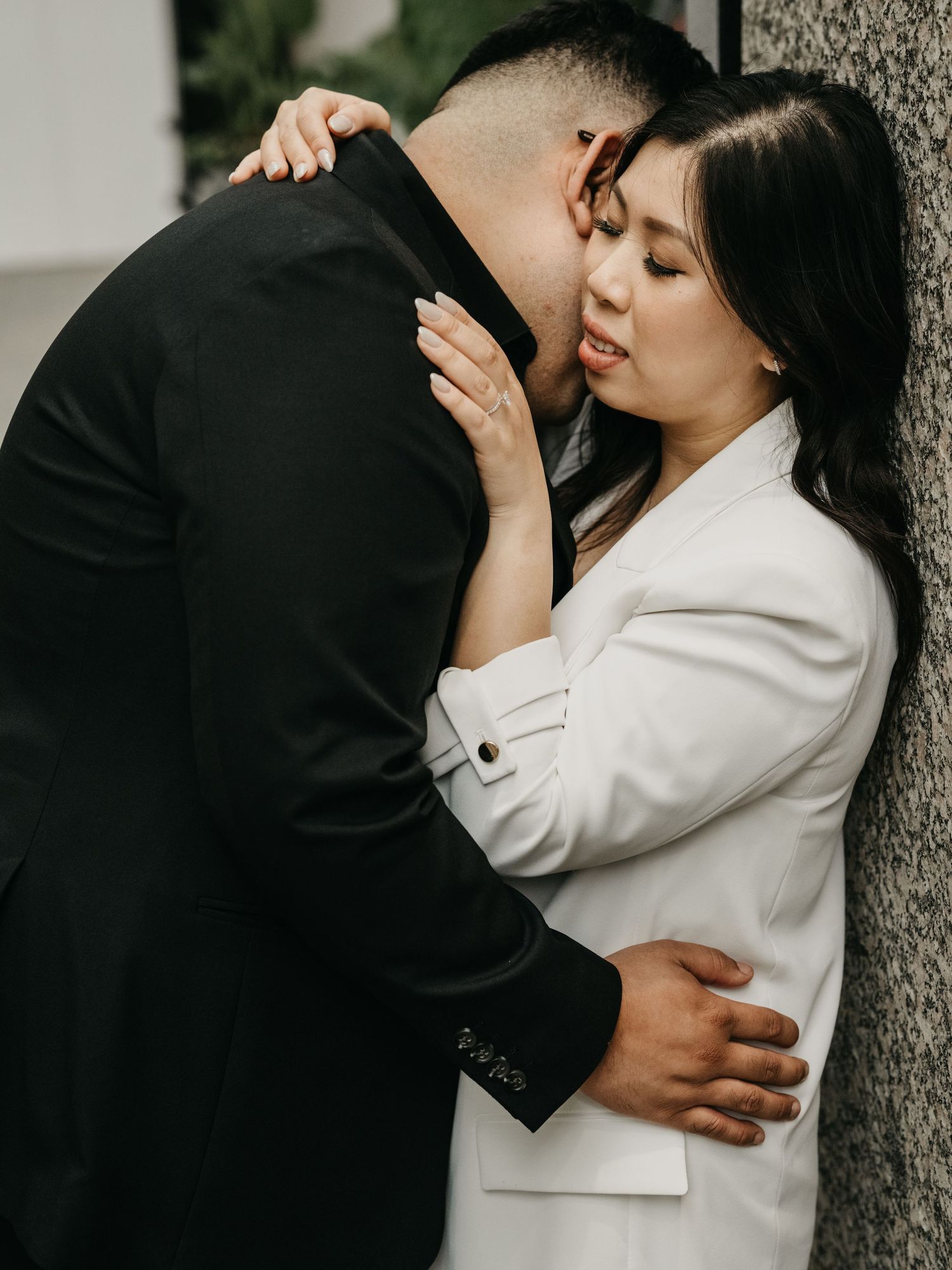 A couple in formal attire shares an intimate embrace against a textured wall in a romantic outdoor photoshoot.