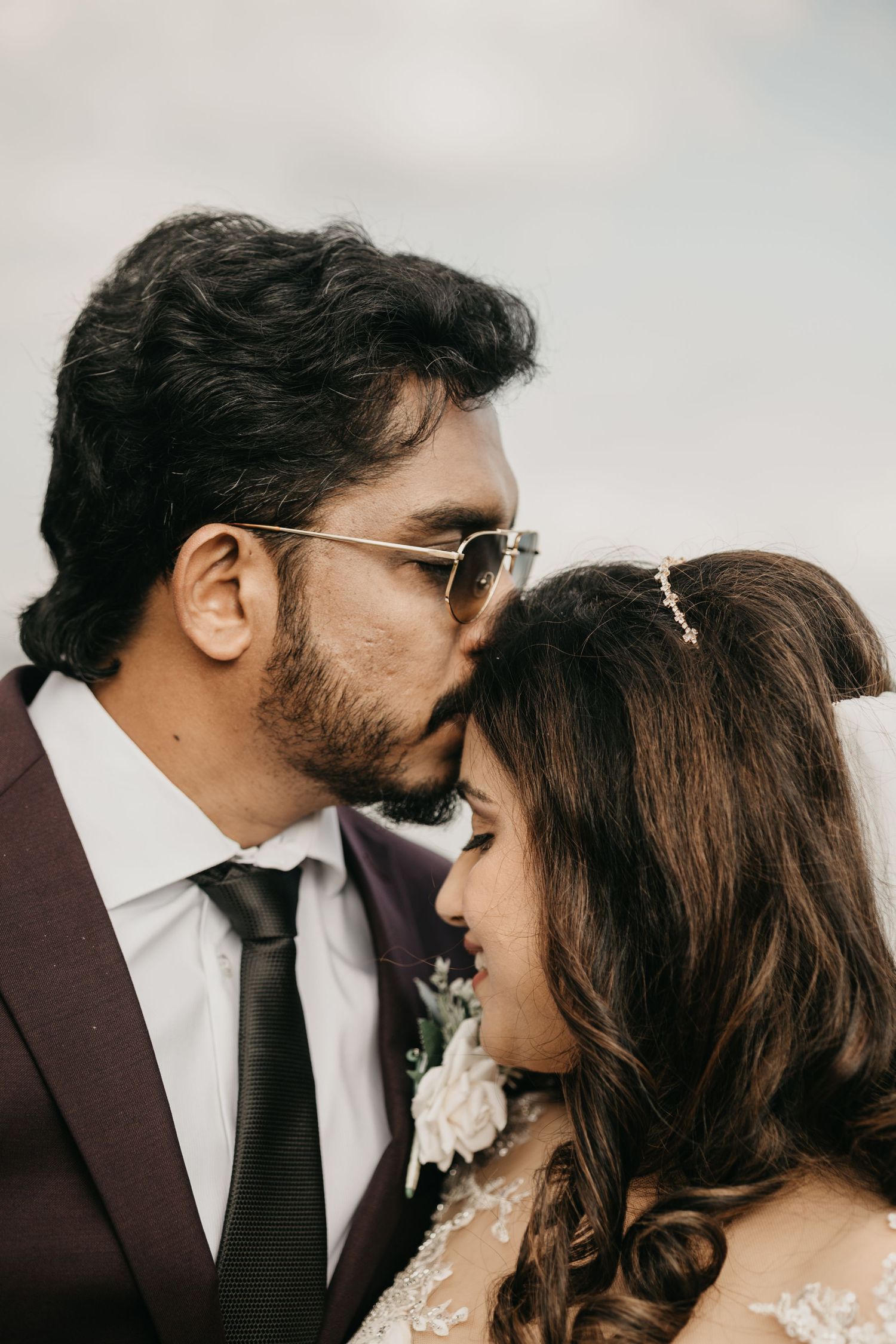 Romantic newlywed couple embrace in intimate wedding portrait against soft cloudy backdrop.