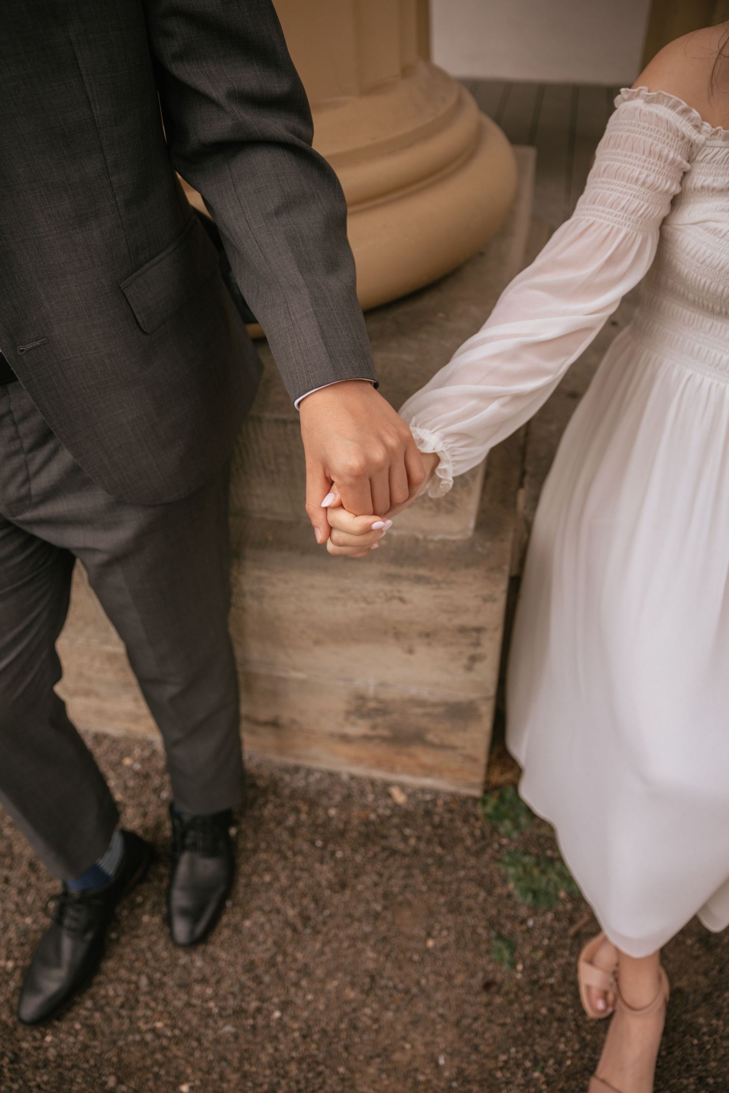 A bride and groom hold hands while standing together, showing her white dress and his dark suit against a rustic backdrop.