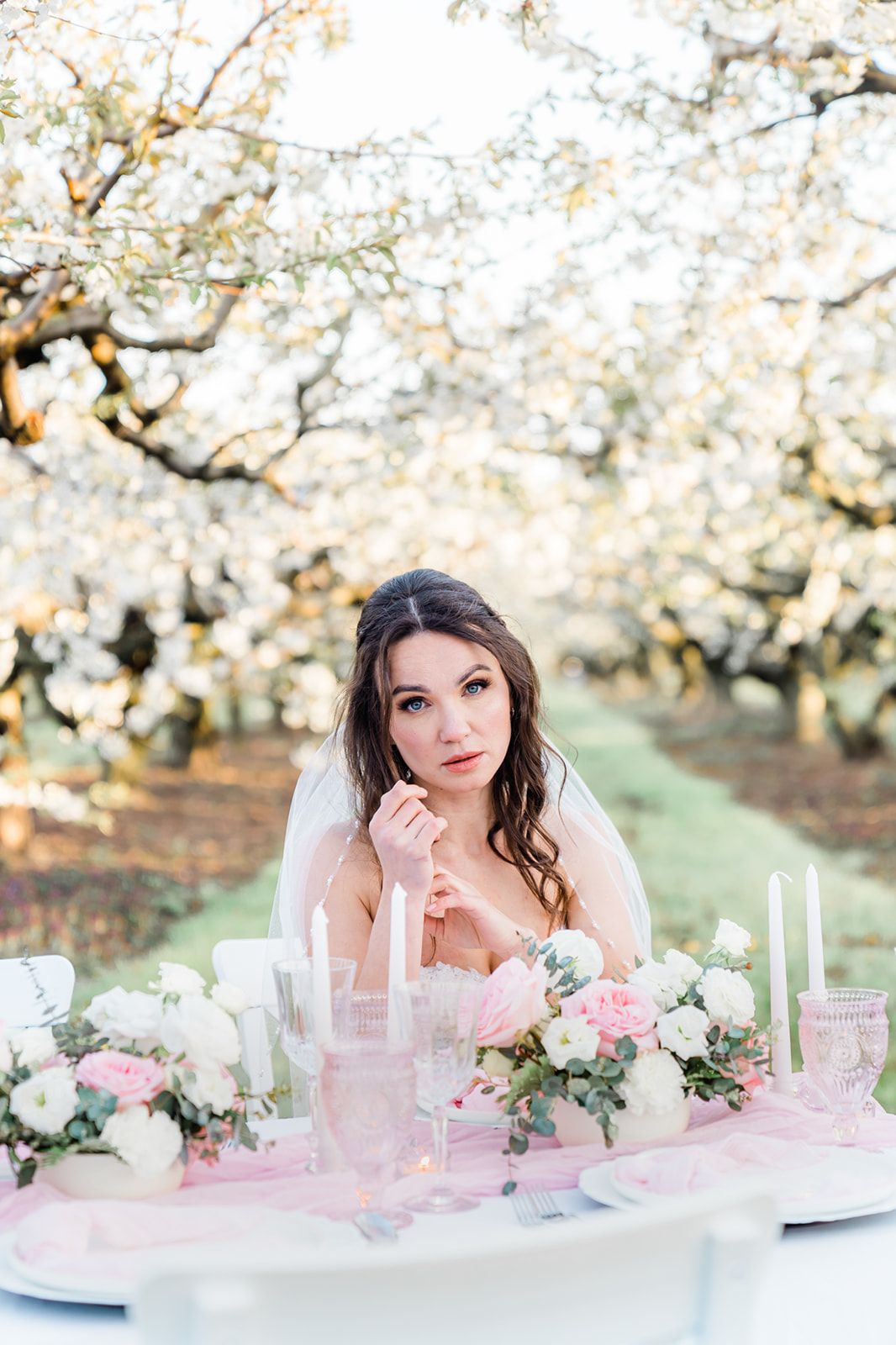 A romantic outdoor table setting with floral arrangements, white candles and pink accents under blooming spring trees.