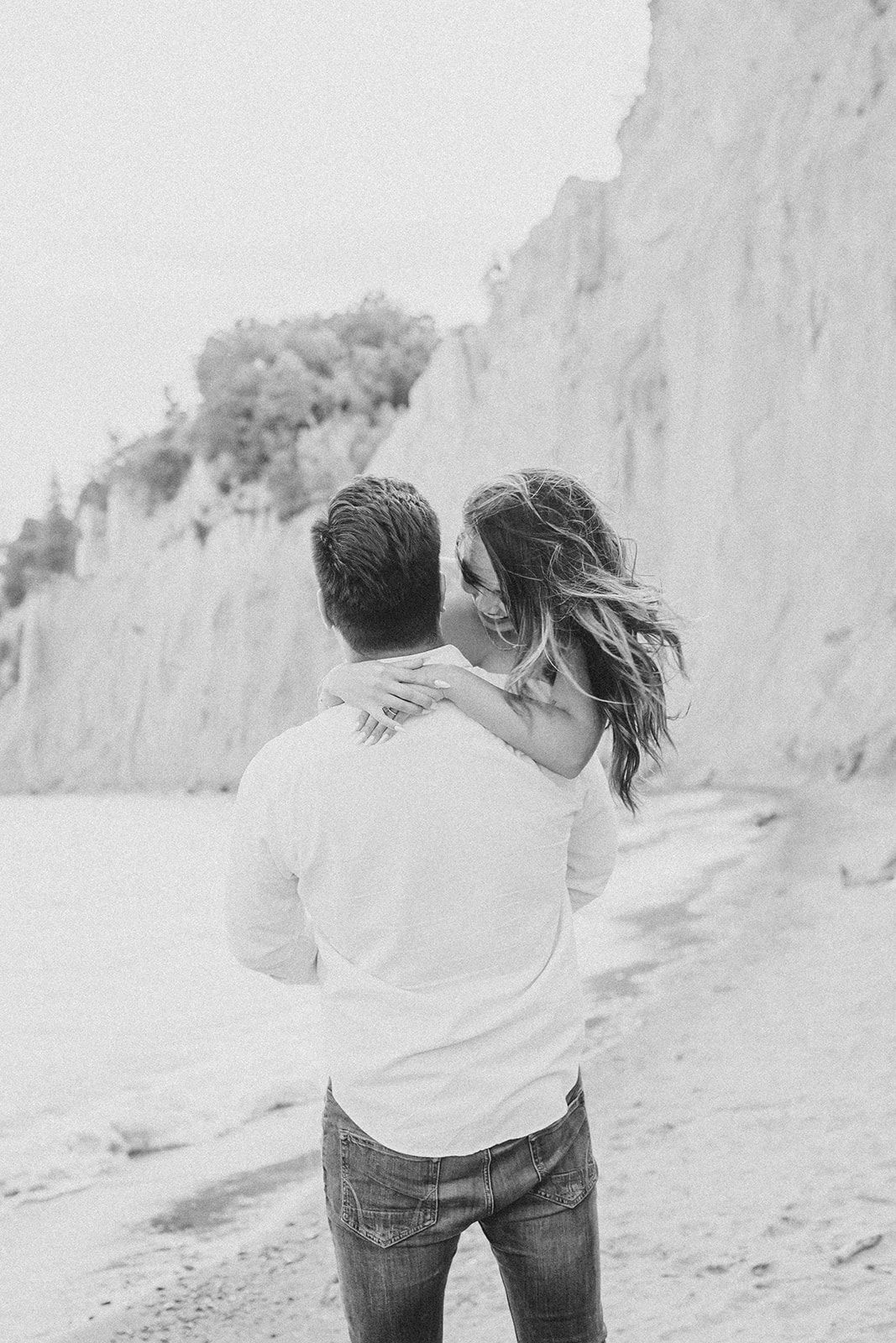 A romantic black and white photo of a couple embracing near dramatic white cliffs on a beach.