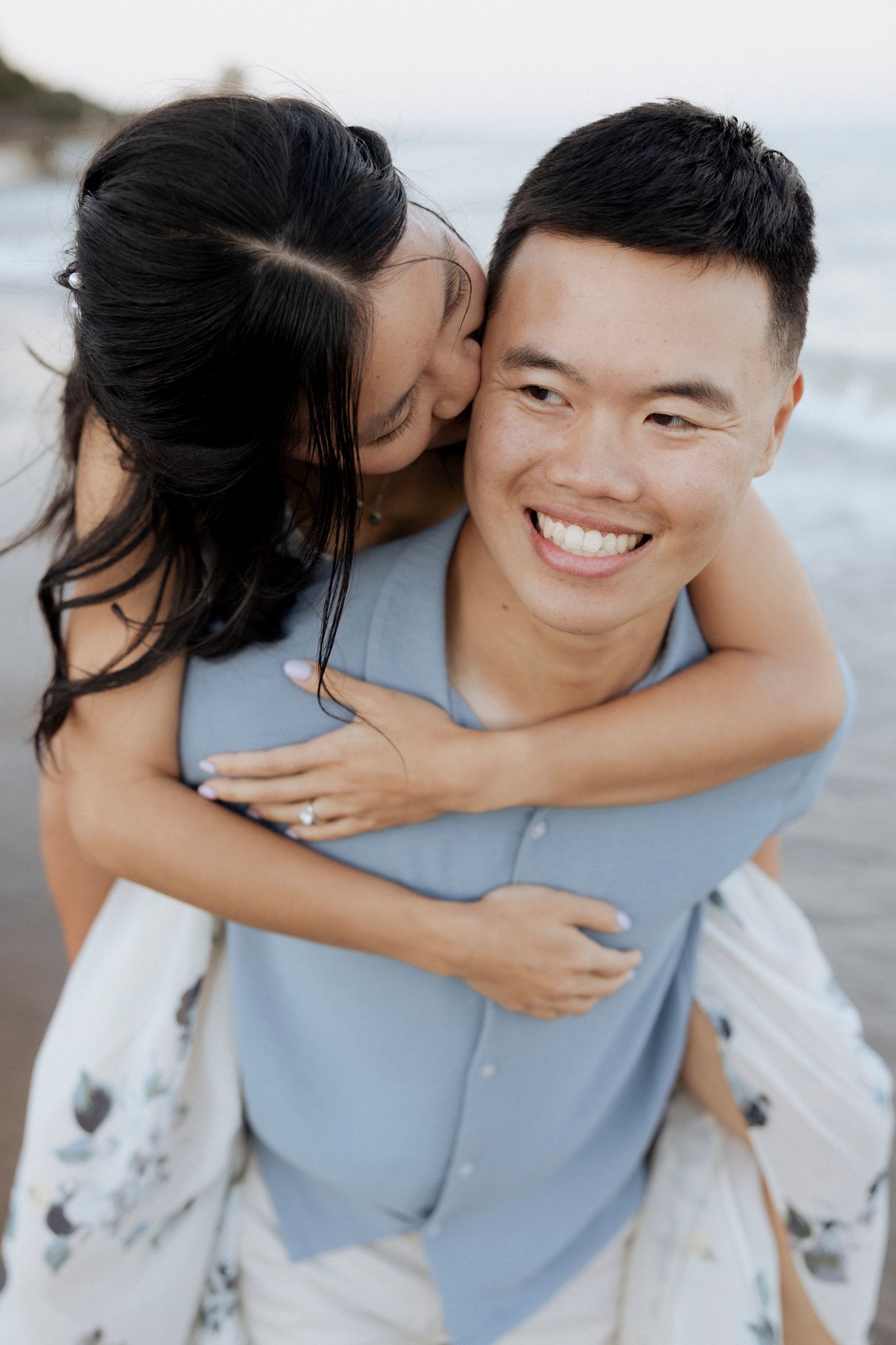 A couple shares a playful piggyback moment at the beach with ocean waves in the background.