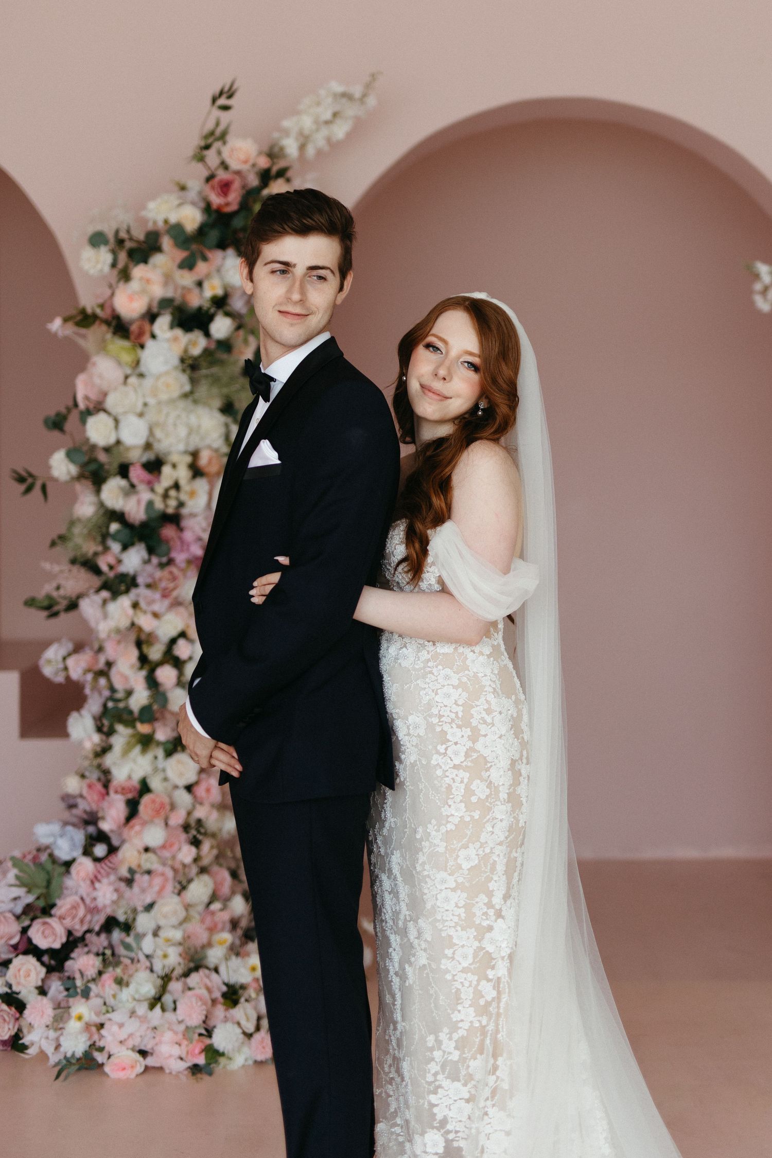 Bride and groom pose together by pink floral arch during wedding ceremony with soft rose colored backdrop.
