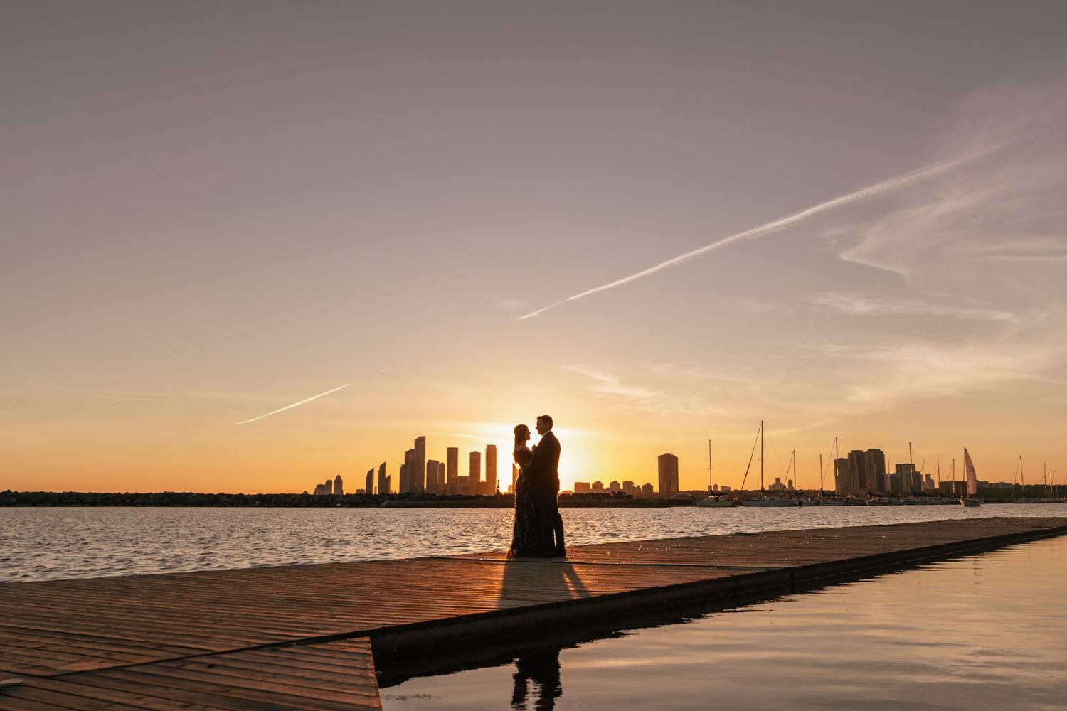 Silhouettes stand on a dock at sunset with city skyline and water reflecting golden hour light.