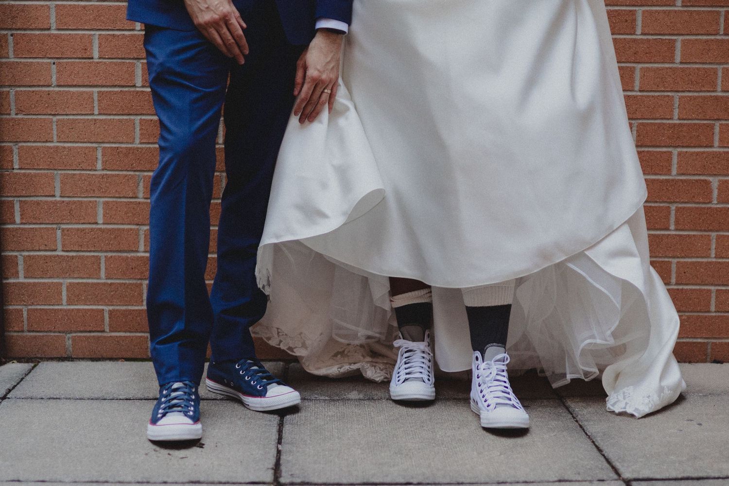 A wedding couple wearing matching white Converse sneakers stands against a brick wall.