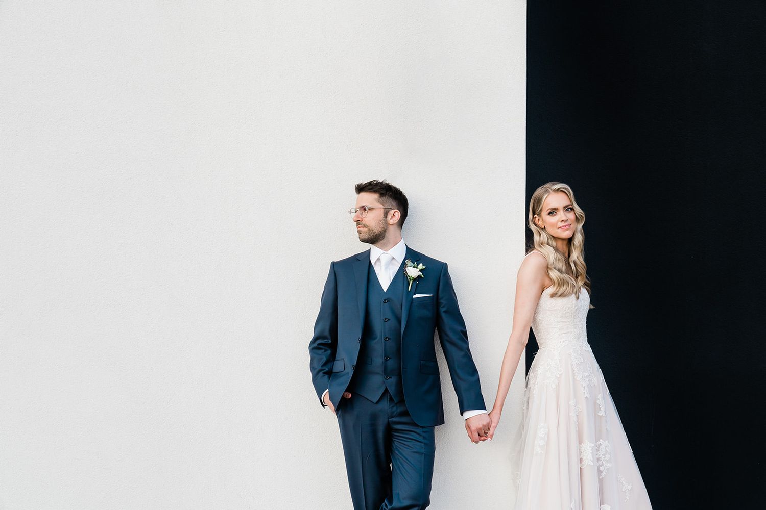 A bride in a white gown and groom in a navy suit hold hands while standing against a black and white backdrop.