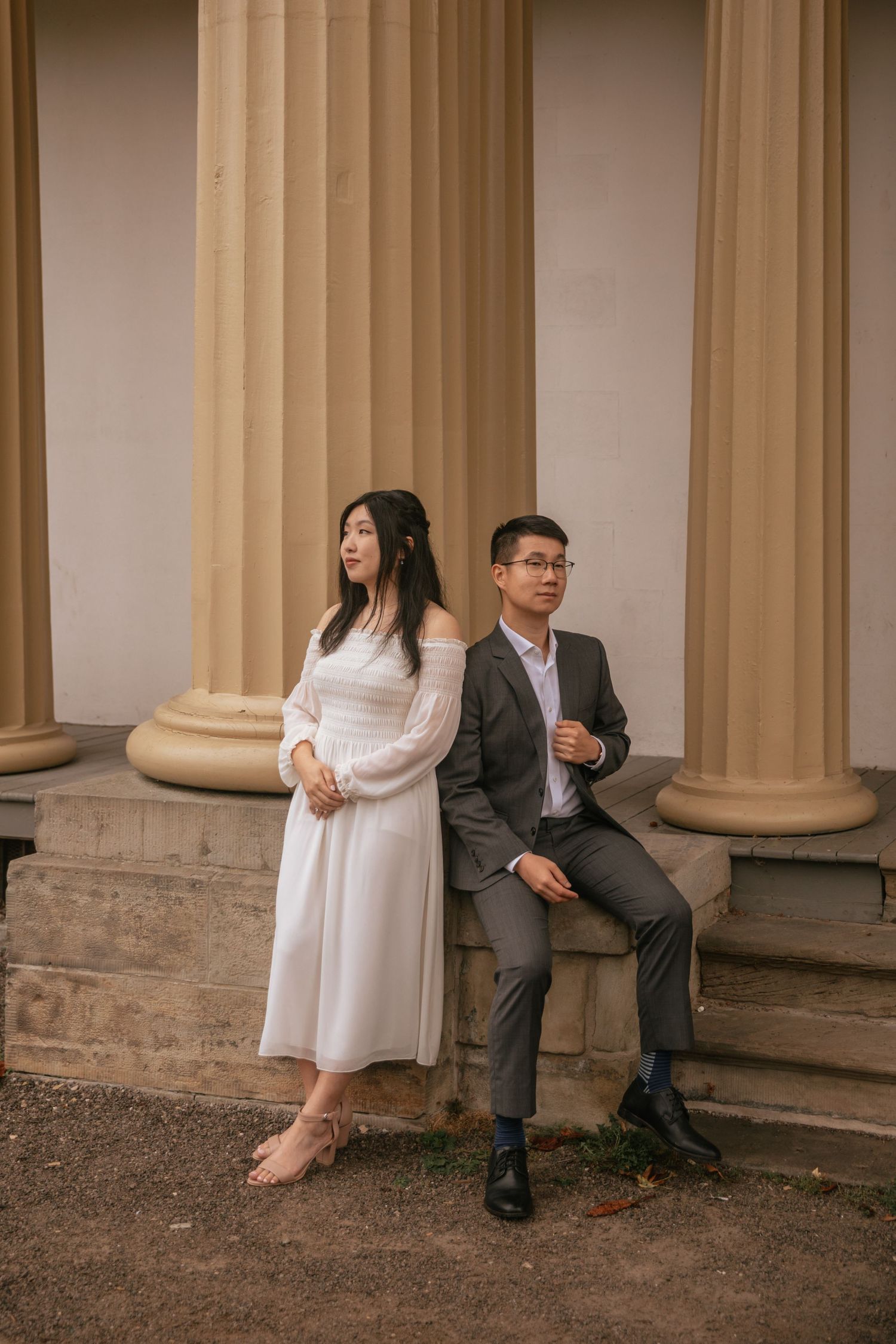 Stylish couple in formal attire poses creatively against classical stone columns at a historic building.